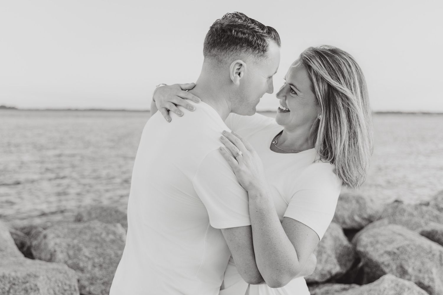 Couple embracing and smiling at each other on rocky beach in black and white engagement photo.