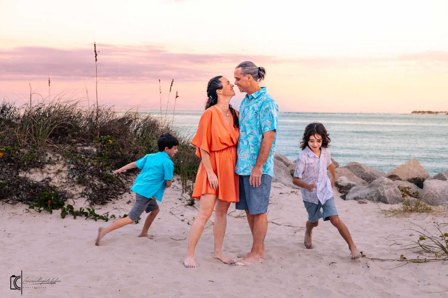 Family in coordinating blue and orange outfits walking together on a sandy beach at sunset.