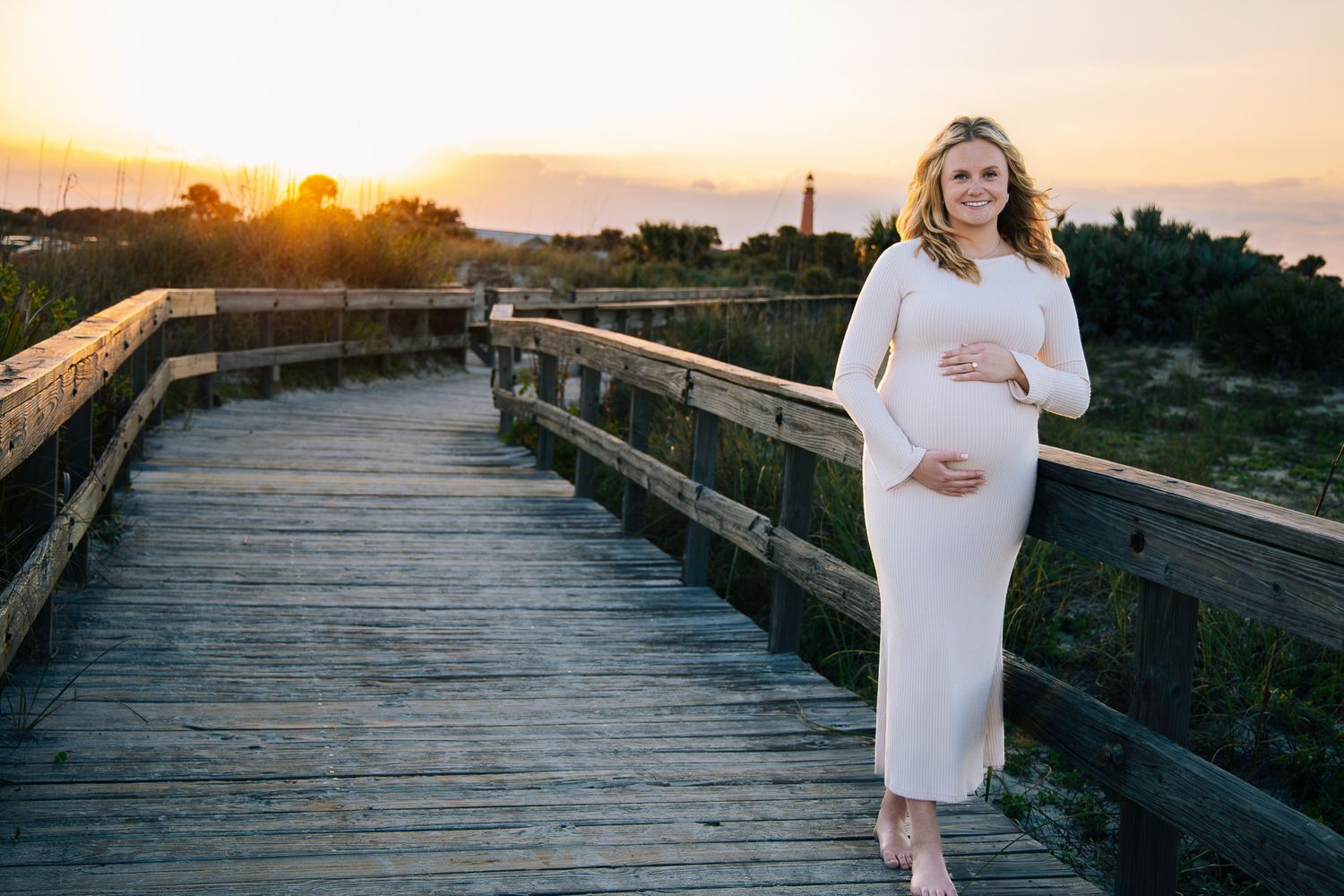 Woman in white dress walks along wooden boardwalk at sunset by the ocean.