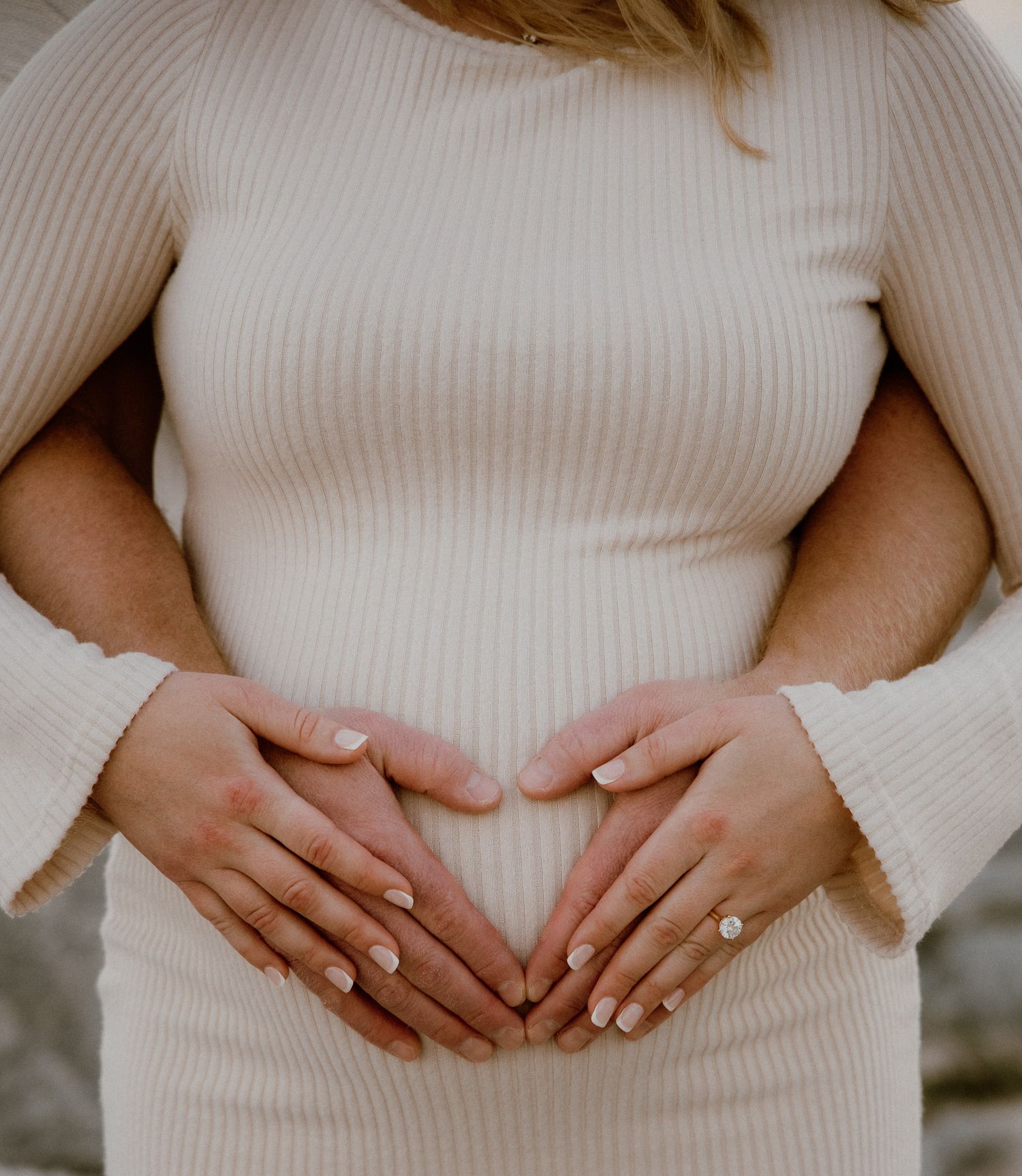 A person wearing a white sweater forms a heart shape with their hands over their stomach in a maternity photo series.