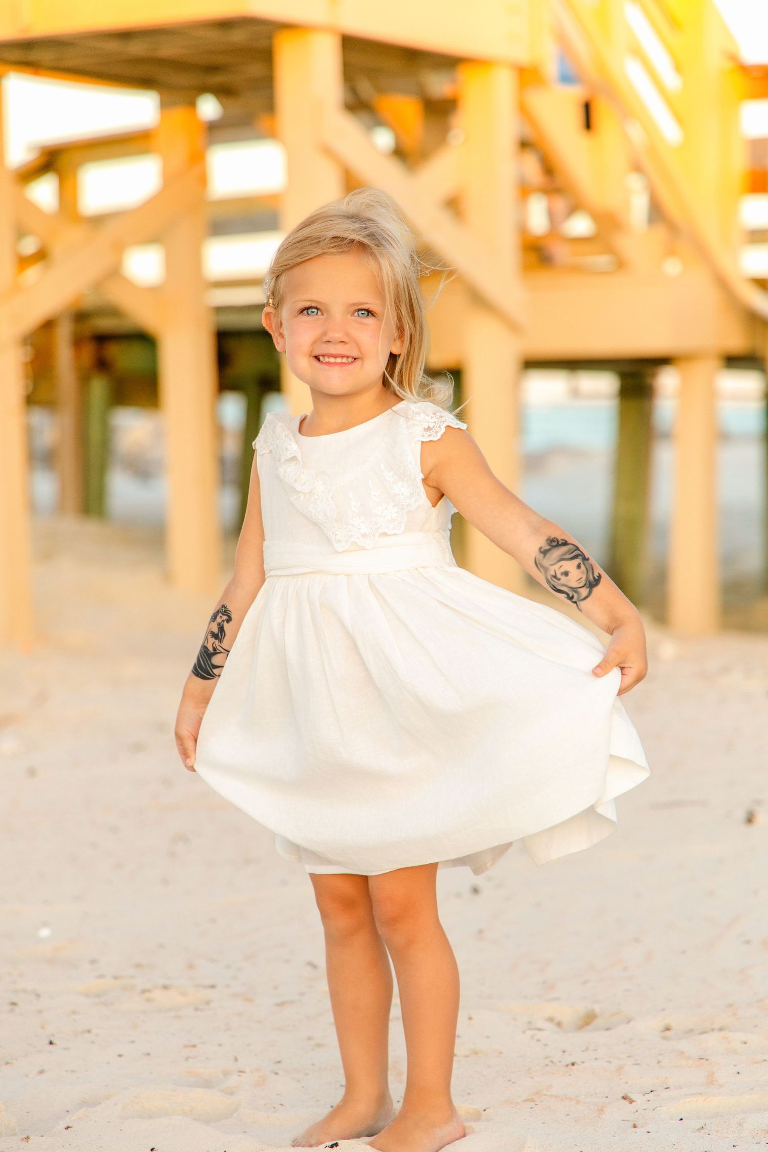Smiling blonde girl in white dress twirling barefoot on sandy beach near pier at golden hour.