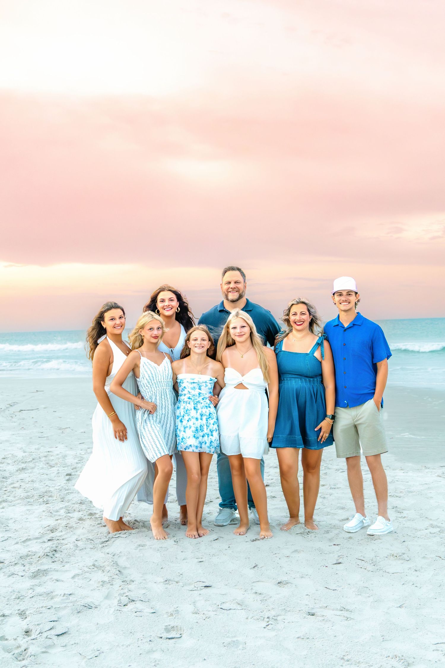 A joyful family of eight poses together on a sandy beach at sunset, dressed in coordinating blue and white outfits.