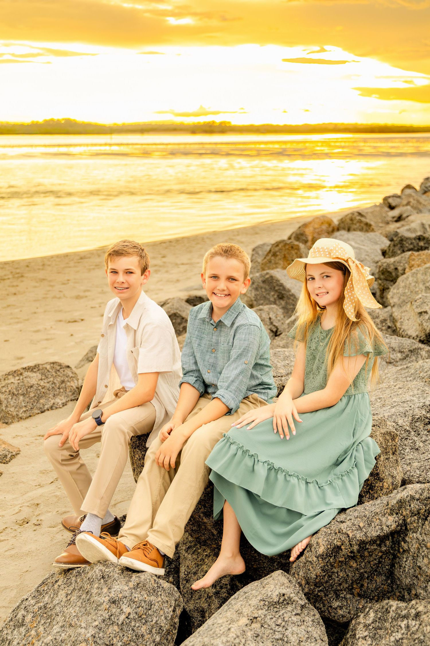 Three children sitting on rocks at a golden sunset beach, two boys in button-up shirts and a girl in a teal dress.