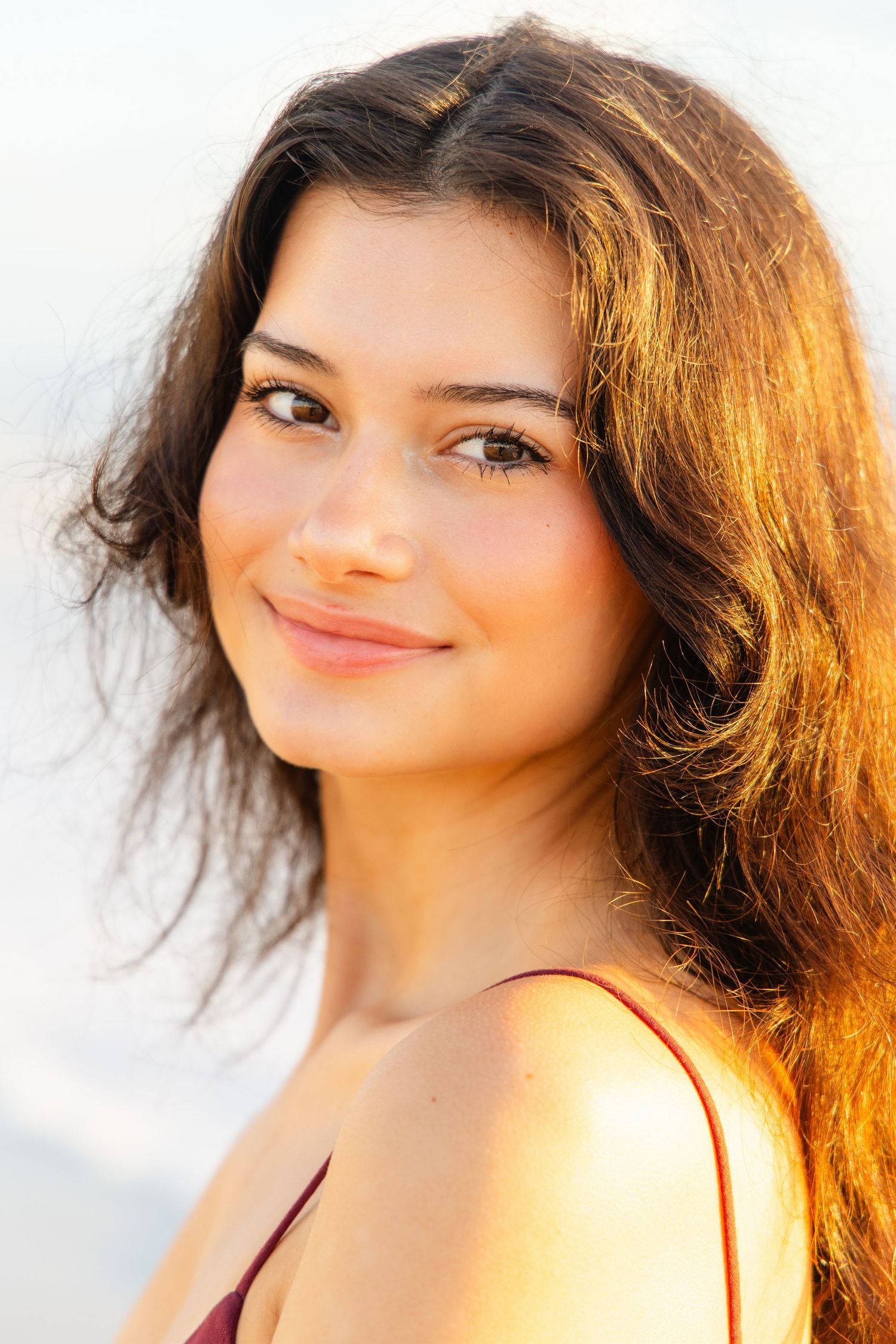 Young woman with dark wavy hair smiling warmly in golden sunset light, wearing a burgundy top.
