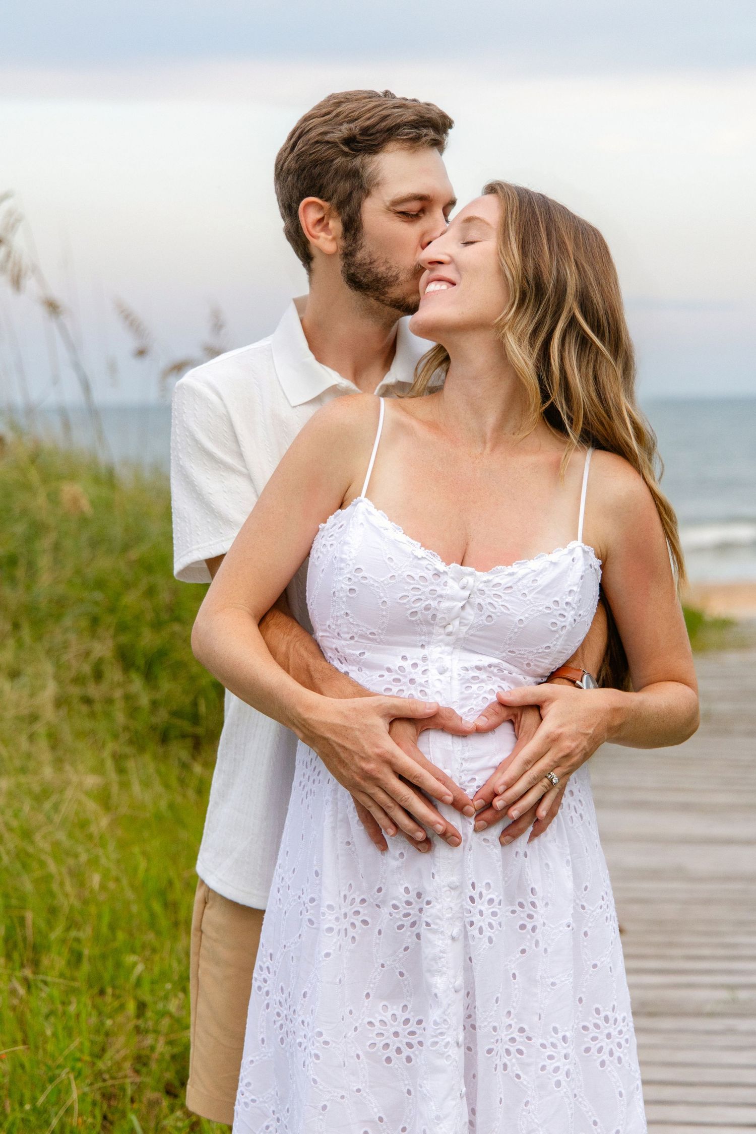 Couple sharing a tender moment on a beach boardwalk, man kissing pregnant woman's temple while forming heart shape on her belly.