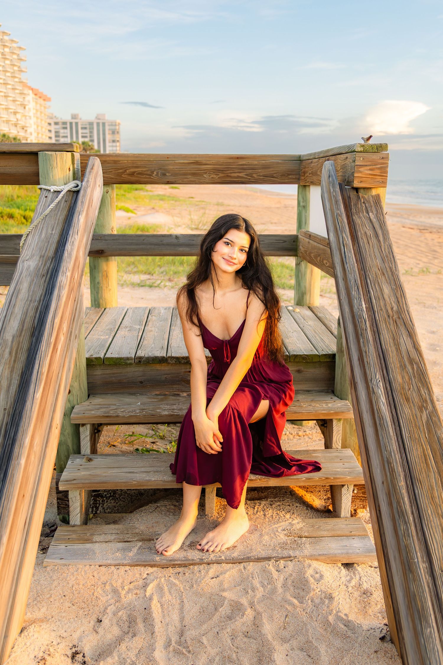 Young woman in red dress sitting on wooden beach stairs, smiling at sunset with ocean and buildings behind her.