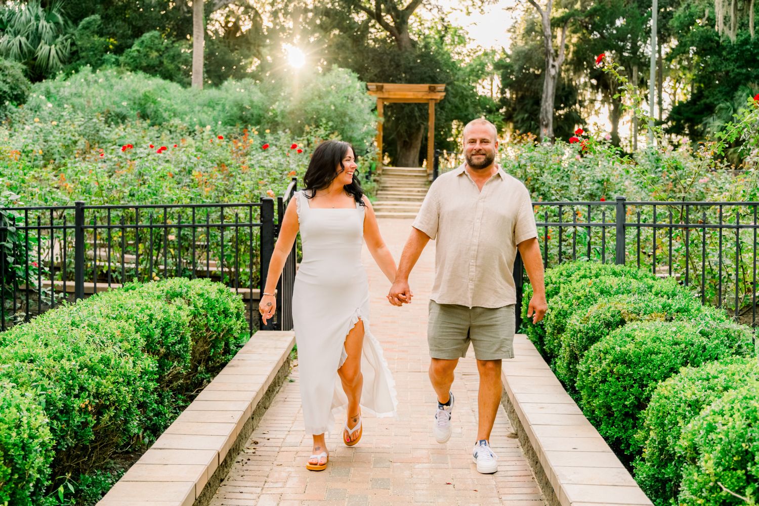 Couple holding hands walking through lush garden pathway with golden sunset light and green hedges.