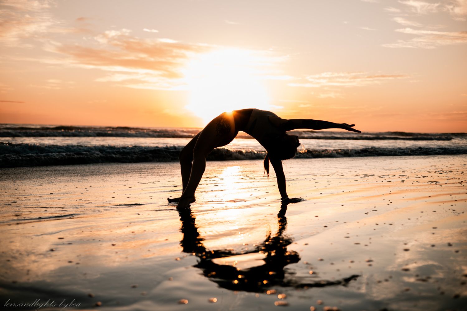 A silhouetted figure performs a backbend on a beach at sunset creating a reflection in the wet sand.