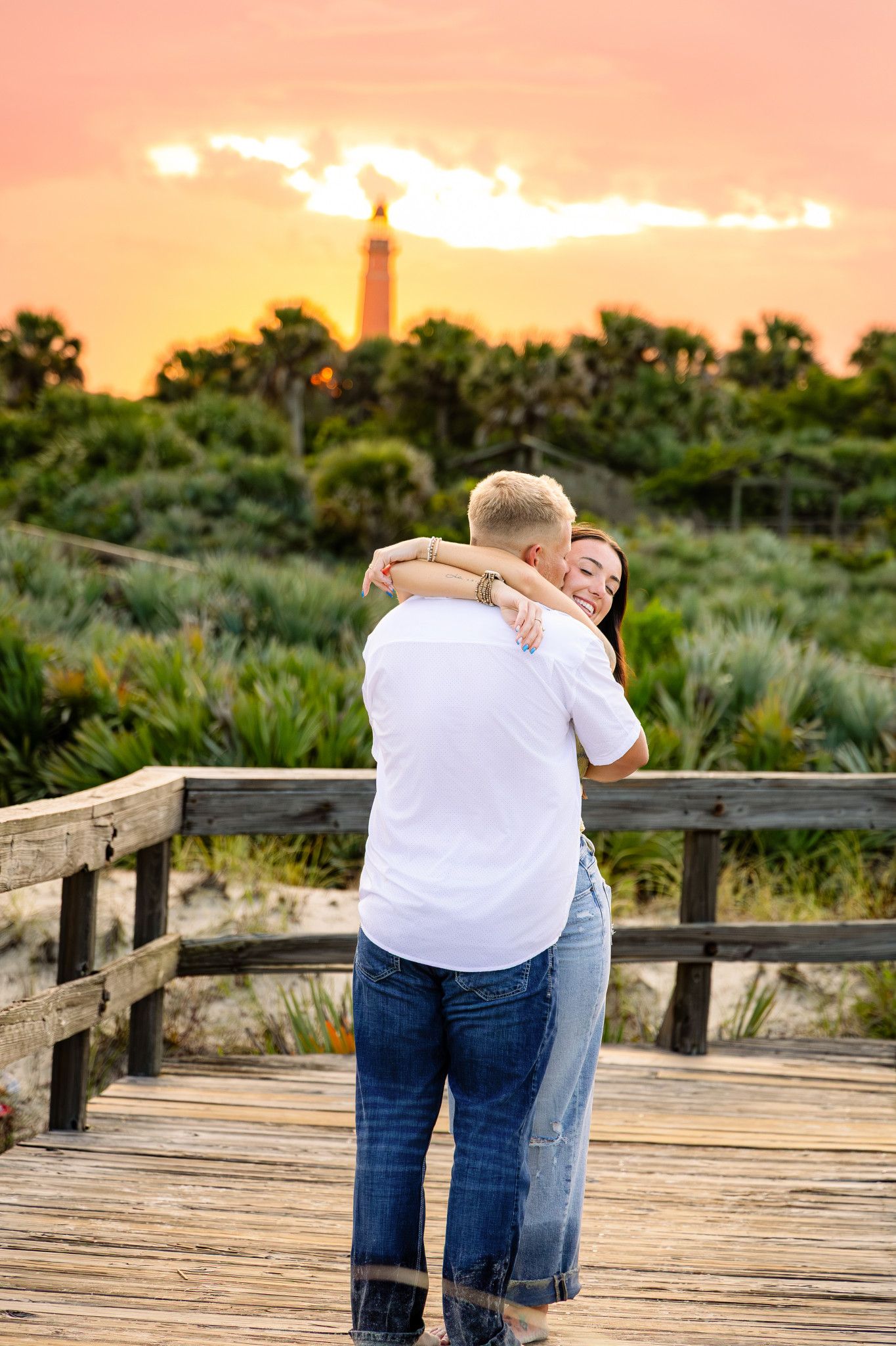 Couple hugging on wooden boardwalk at sunset with lighthouse and palm trees visible in background.