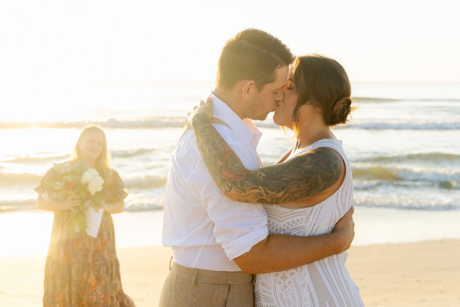 Couple shares romantic kiss during beach wedding ceremony with ocean waves in background.