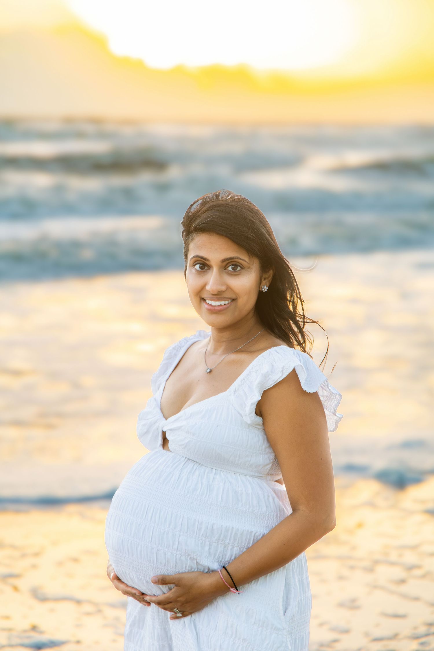 Smiling pregnant woman in a white dress cradling her belly on a sandy beach at golden sunset for a maternity photo.