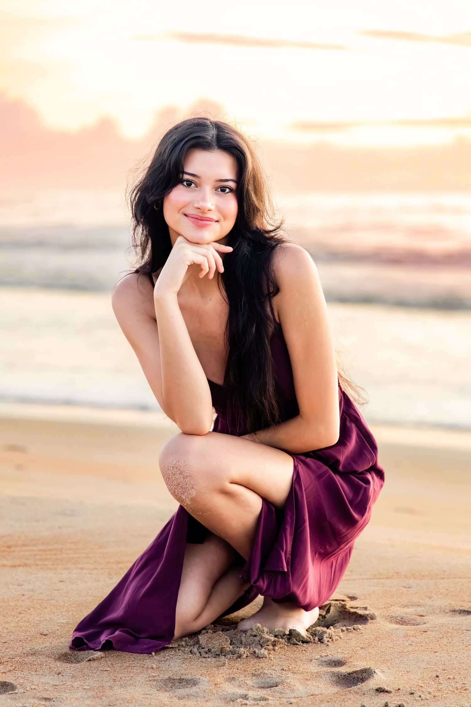 Young woman in purple dress crouching on sandy beach at sunset, smiling with hand on chin.
