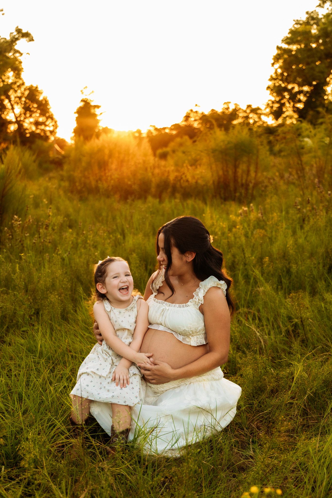 Pregnant woman in white dress sits in golden sunset field with laughing toddler girl touching her baby bump.