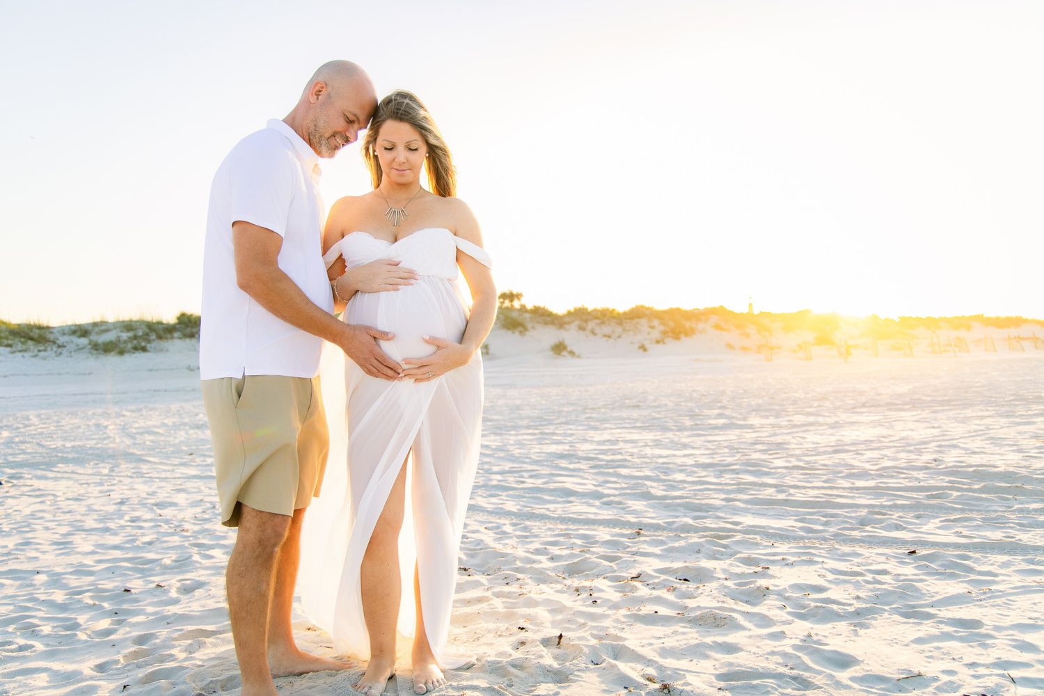 Pregnant couple embracing on a sunny beach at golden hour, woman in white maternity dress cradling her baby bump.