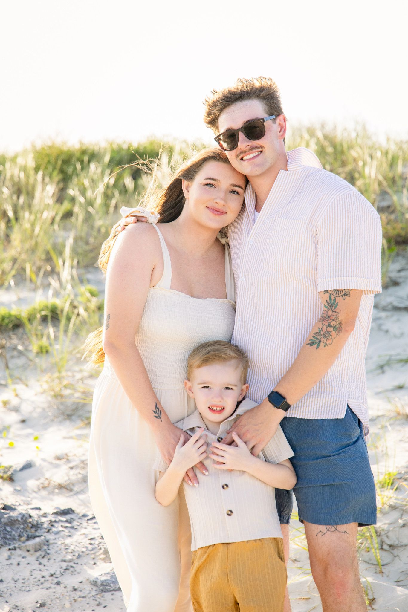 Family of three posing together on sandy beach dunes, parents embracing young toddler boy in warm golden light.