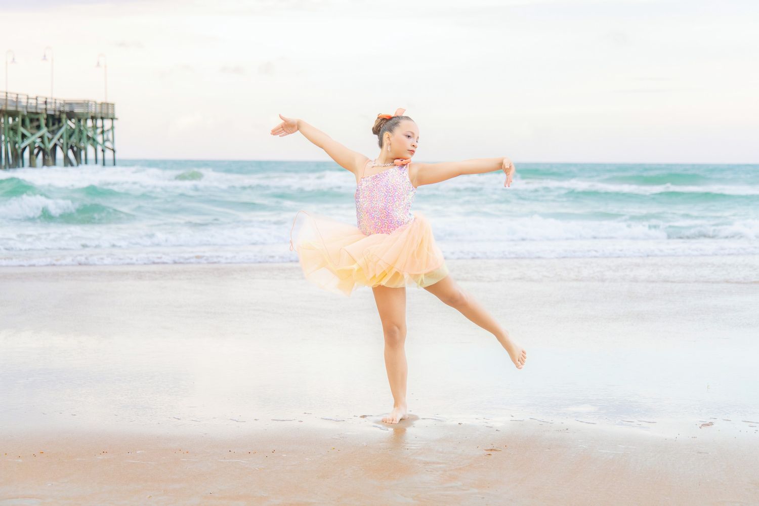 Young girl in pink tutu ballet dress dancing gracefully on a sandy beach with ocean waves in the background.