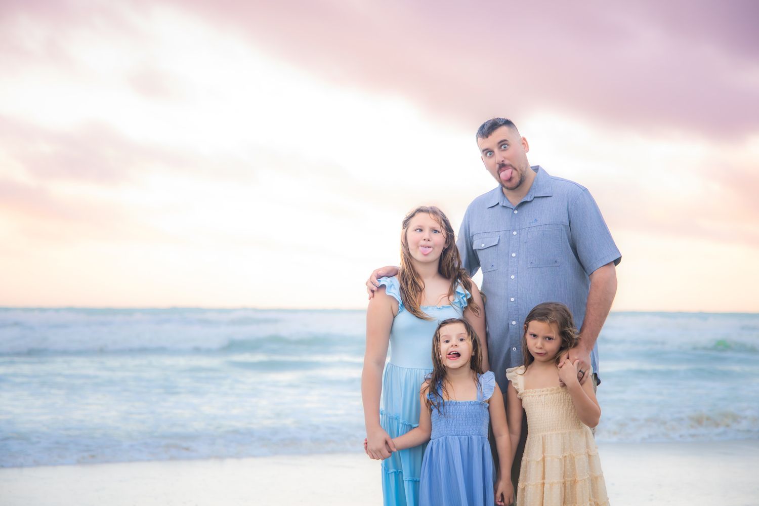 Family of four posing on a sandy beach at sunset with pastel skies and ocean waves in the background.