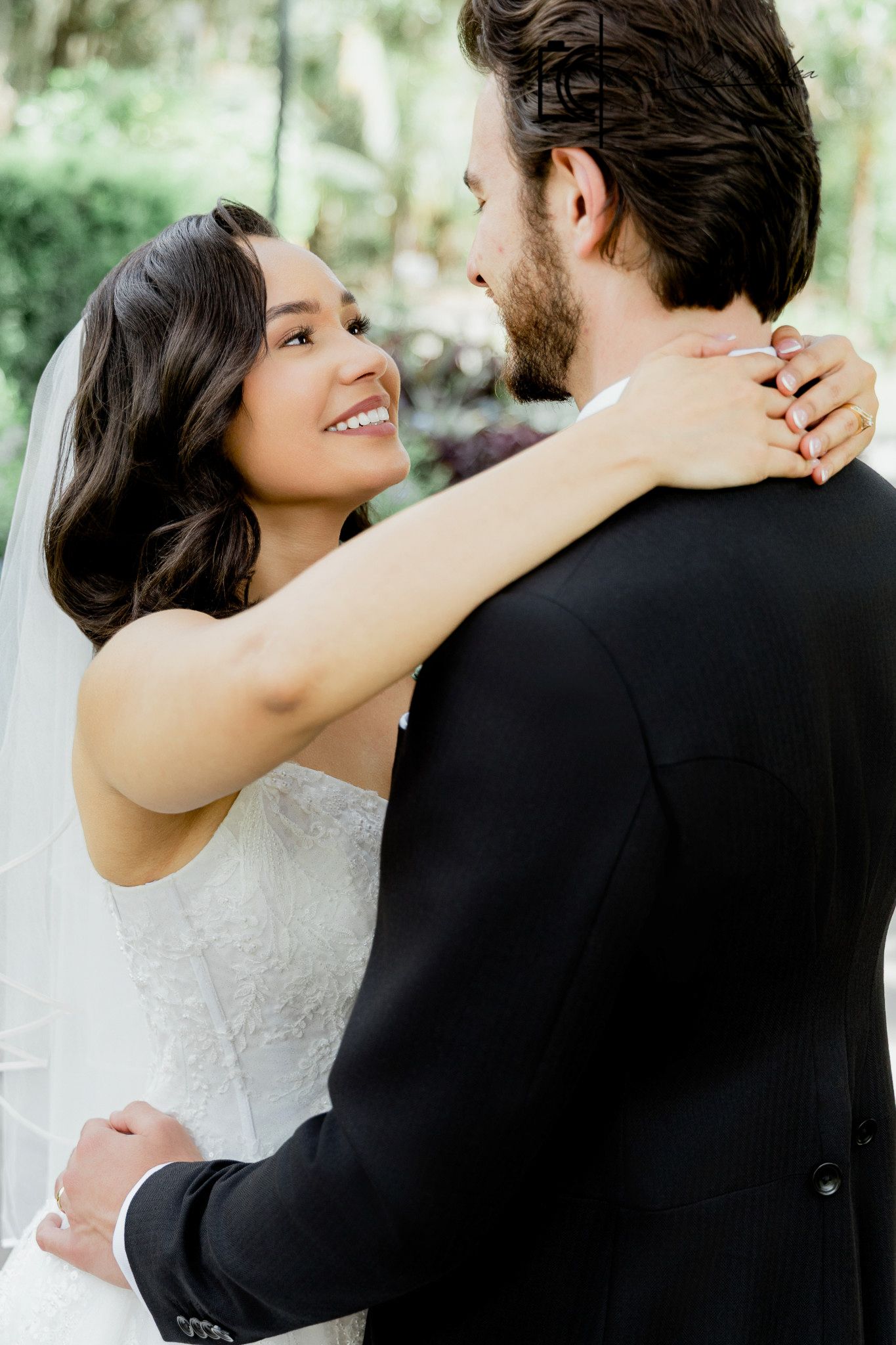 Smiling bride in white lace gown and veil embraces groom in black suit during outdoor wedding ceremony.