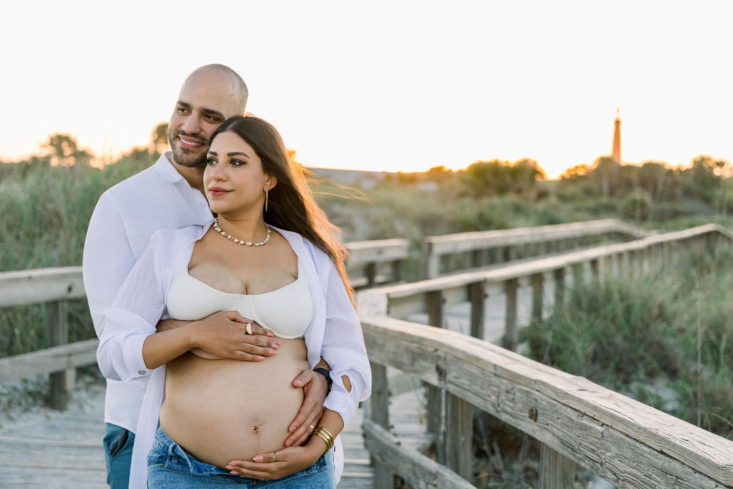 Couple embracing on a boardwalk at sunset, woman showing pregnant belly, golden hour beach backdrop.
