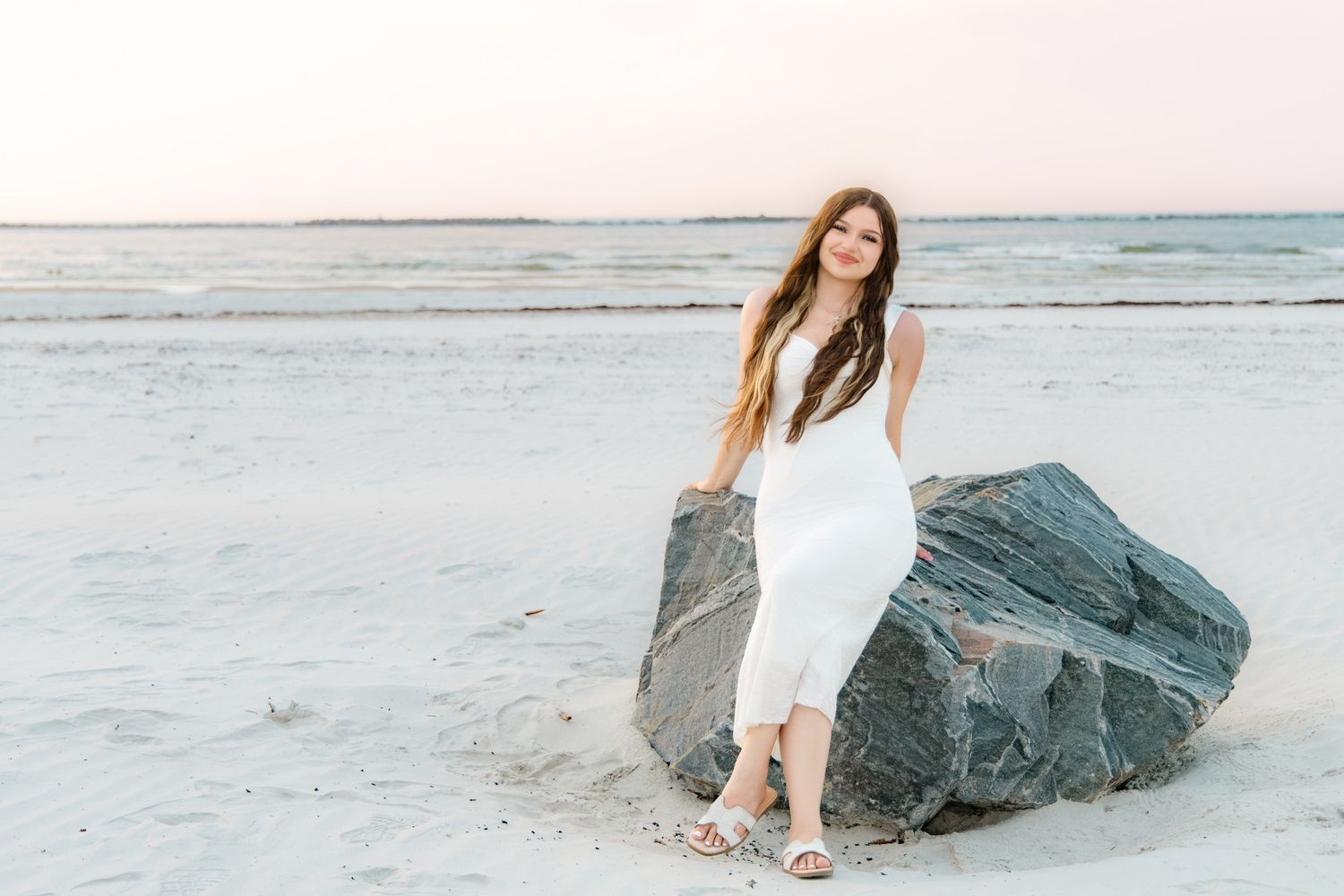 Woman in white dress leaning on large rock on sandy beach at sunset.