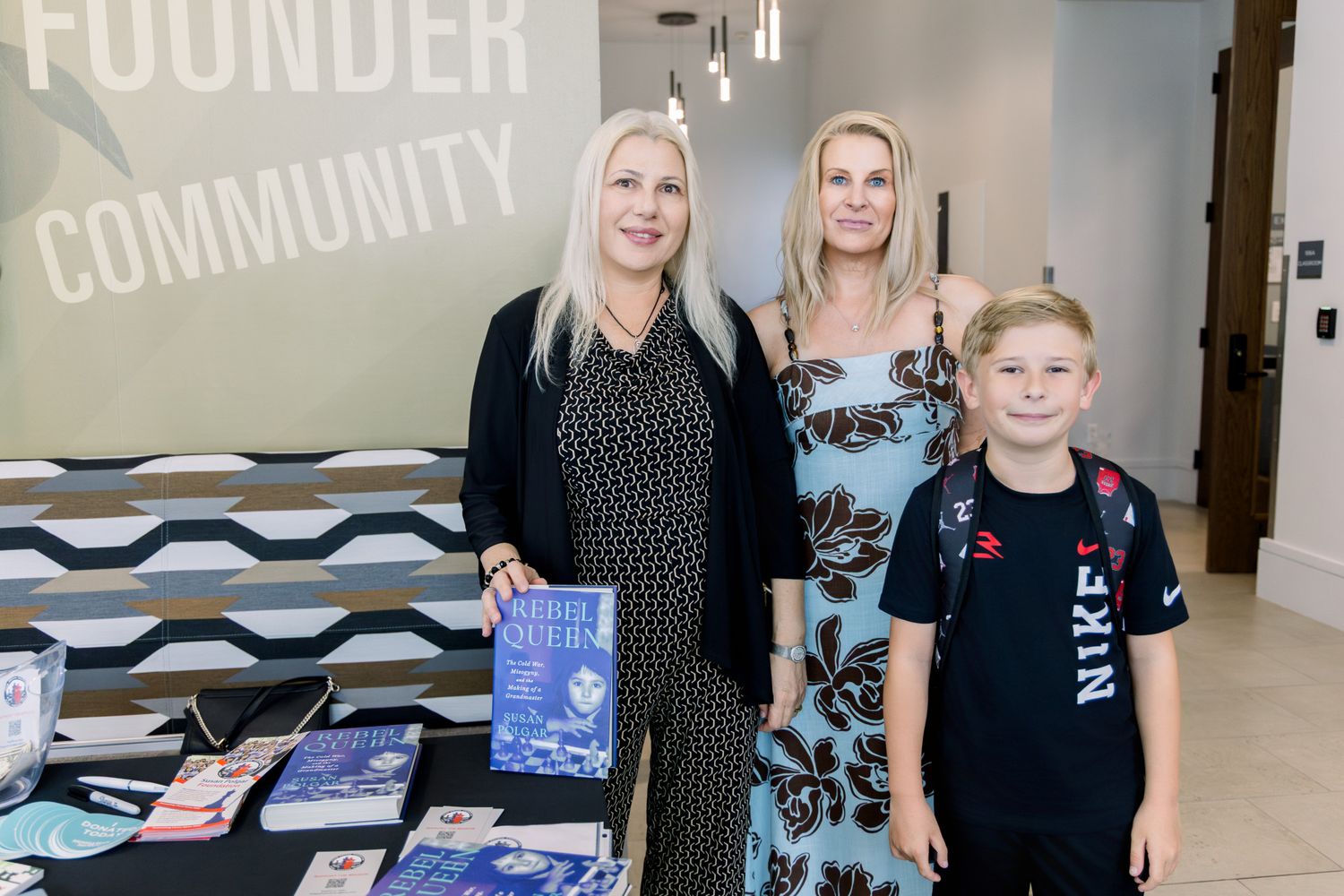 Two women and a young boy pose at a Founder Community book signing event table displaying books.