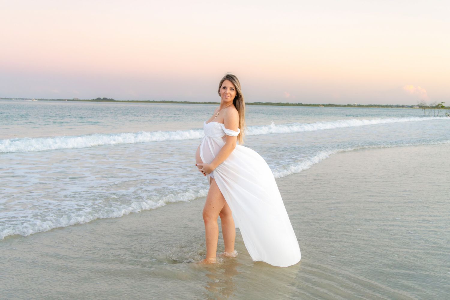 Pregnant woman in white off-shoulder gown standing in ocean waves at sunset beach maternity photo session.