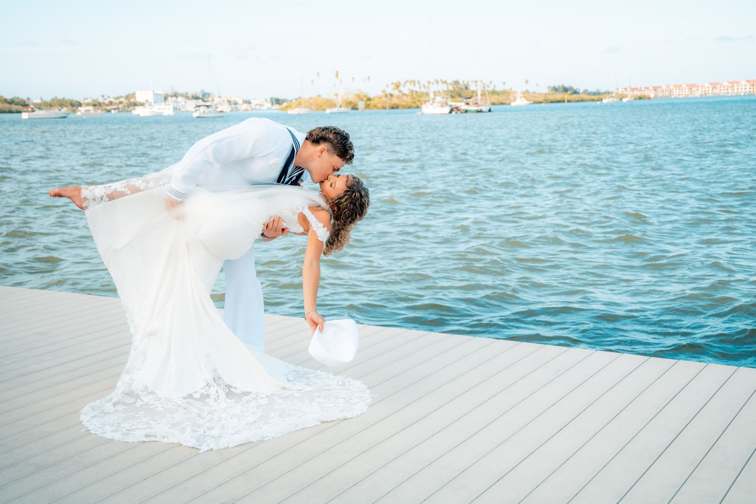 Bride and groom sharing romantic dip kiss on waterfront dock in wedding attire with blue water backdrop.