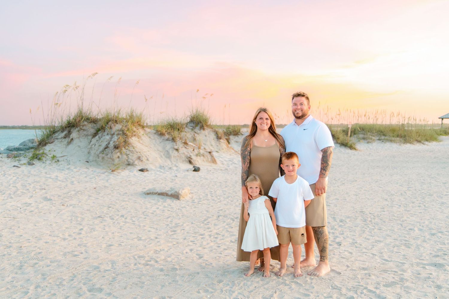Family of four poses on a white sand beach at sunset with sea oats and pastel sky backdrop.
