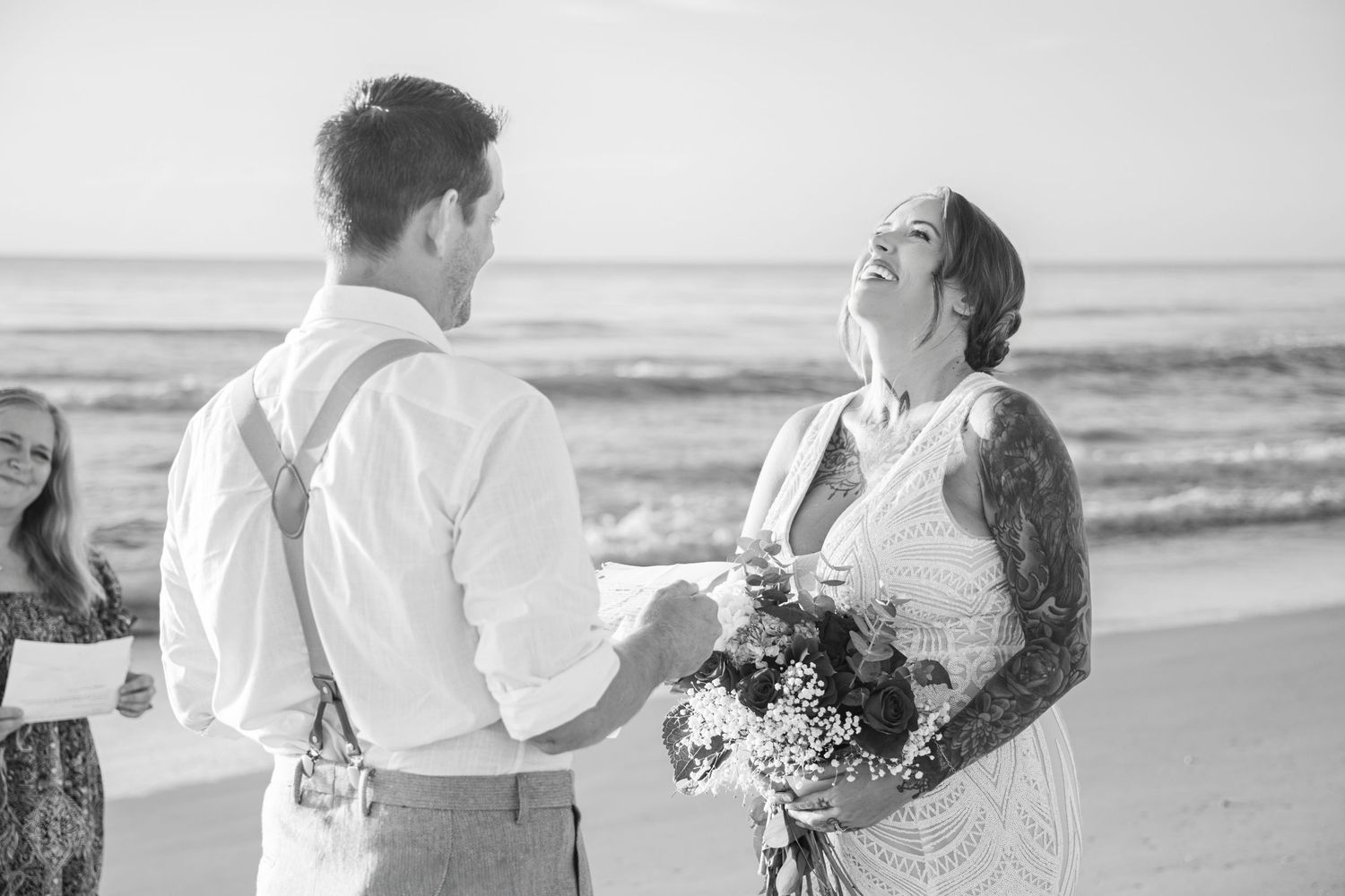 Black and white beach wedding photo of tattooed bride laughing while exchanging vows with groom holding hands.