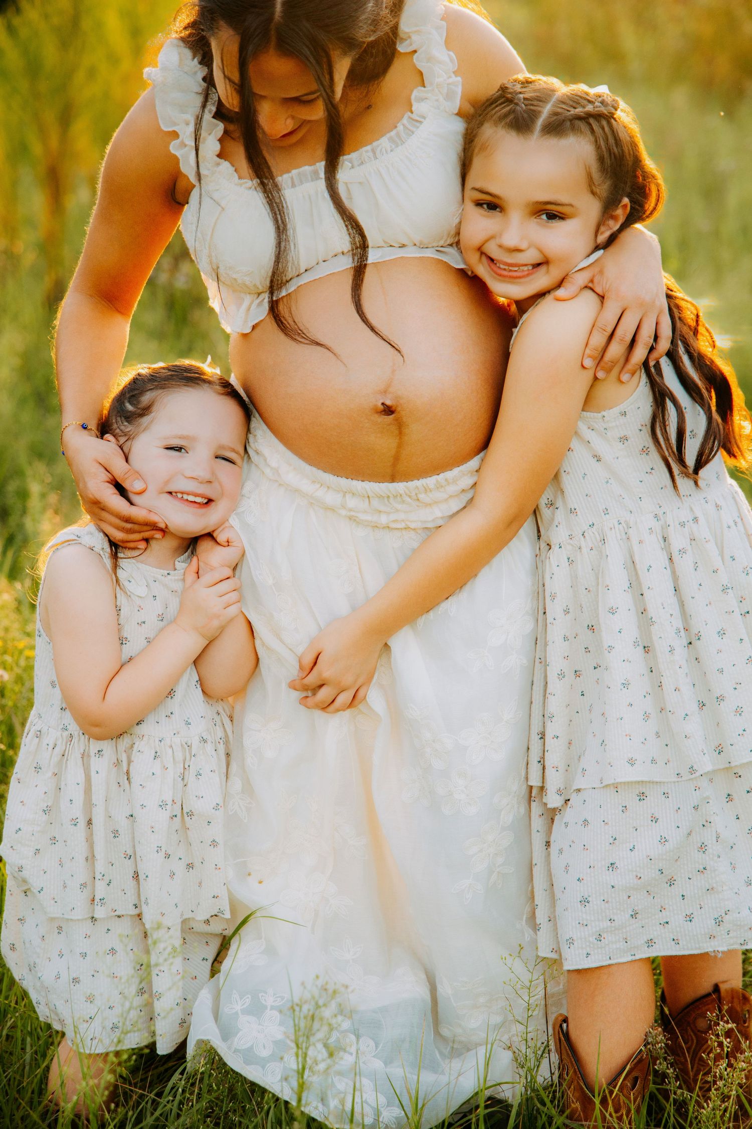 Pregnant woman in white dress outdoors with two young daughters hugging her baby bump in golden sunlight.