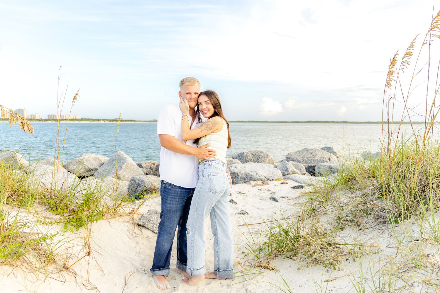 Couple embracing barefoot on a sandy beach near rocky shoreline with calm blue water and sea oats nearby.