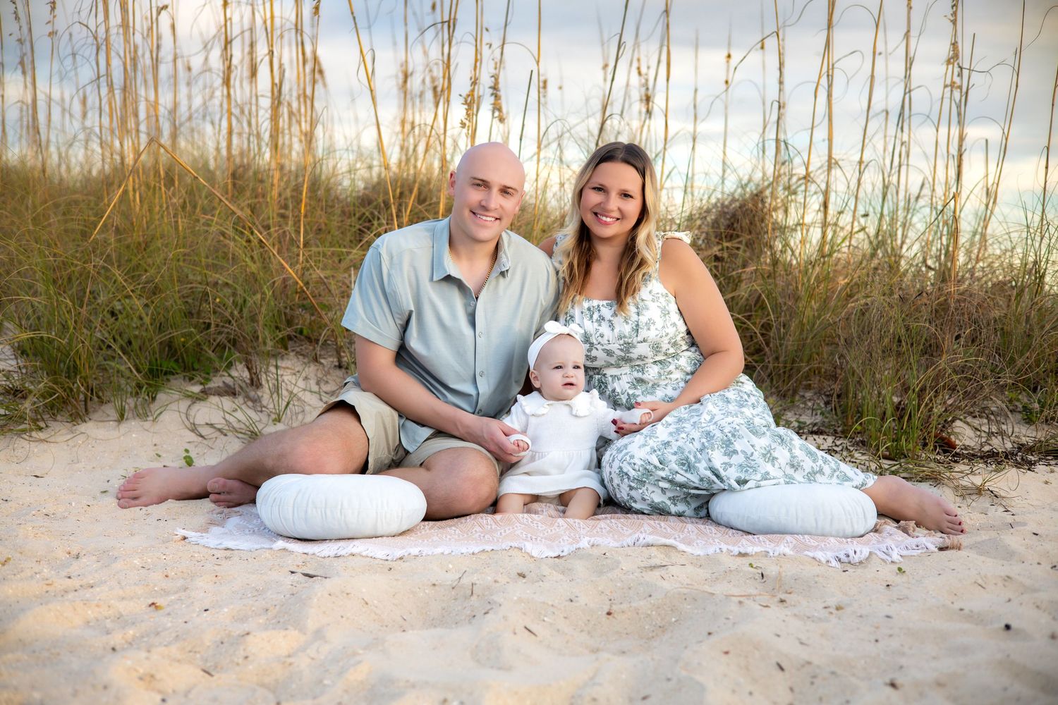 Family group wearing coordinated white and blue outfits relaxing together on a sandy beach at sunset.