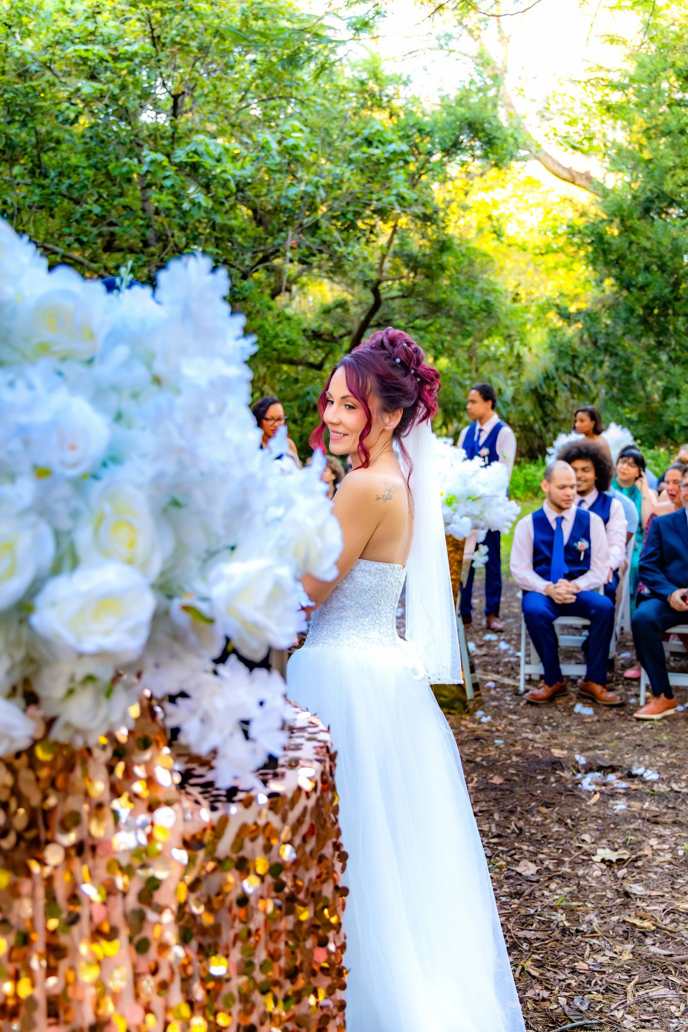 A bride in a white wedding dress poses in a garden setting with guests in blue attire visible in the background.
