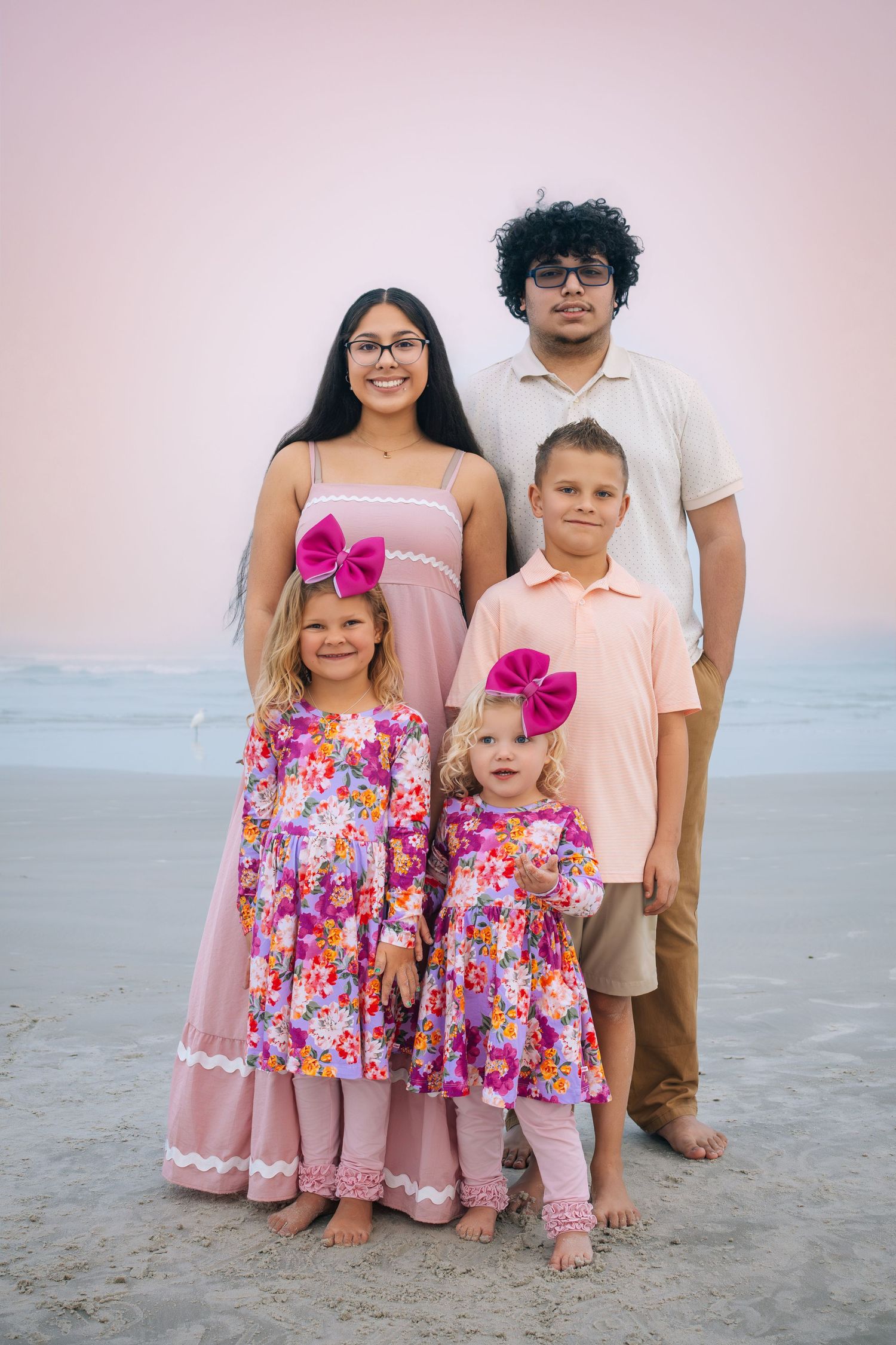 Family wearing coordinated pink outfits poses together on a beach at sunset.