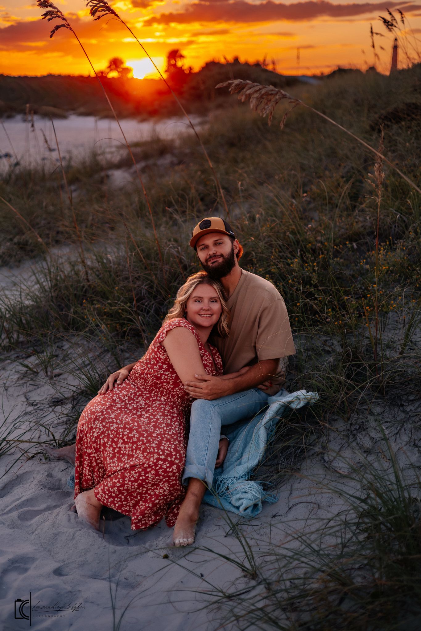 A couple embraces on sandy dunes at sunset with sea oats silhouetted against an orange sky.