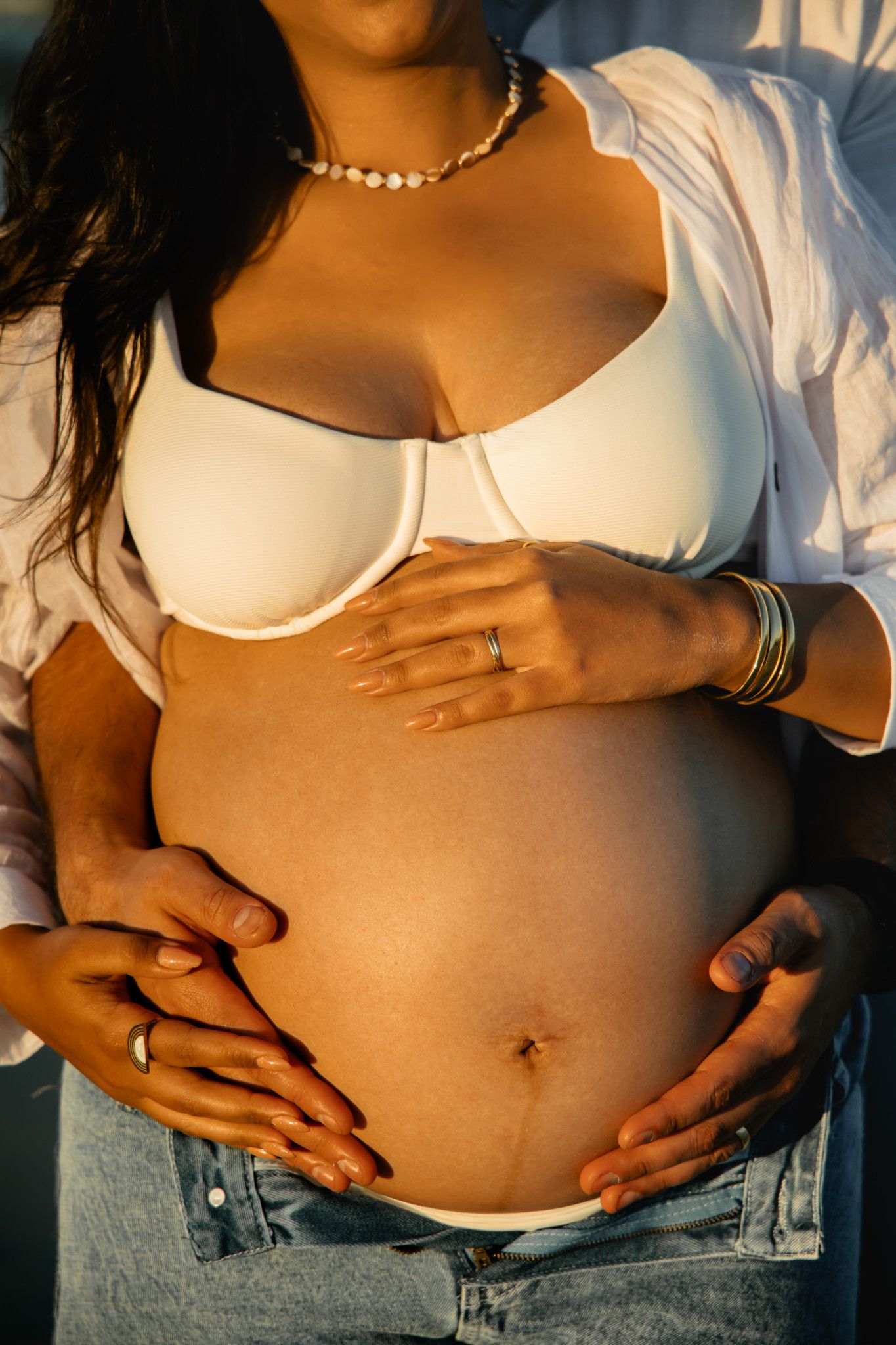 Pregnant woman in white bra and jeans cradling baby bump with partner's hands, golden hour lighting.