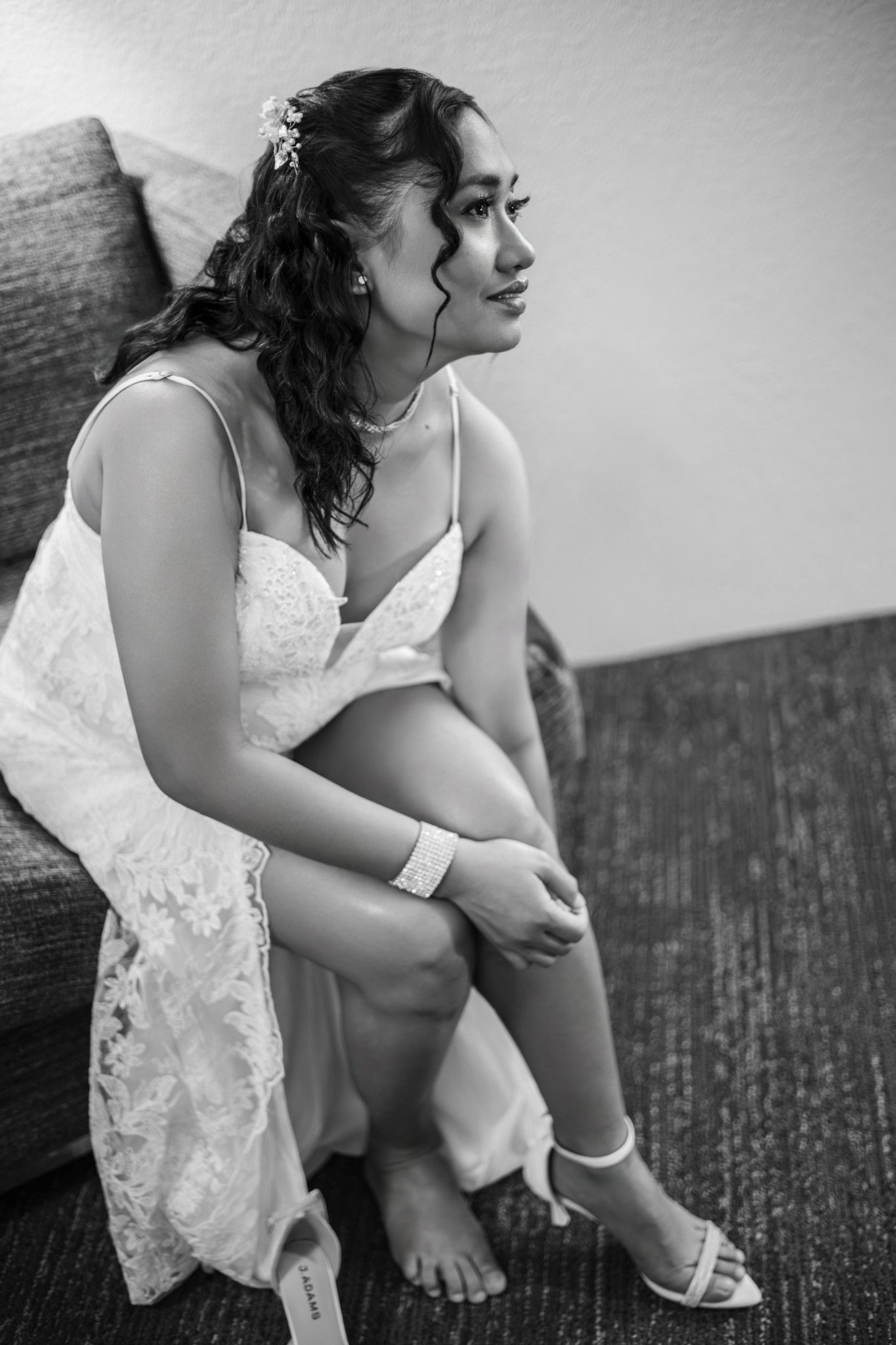 Black and white photo of a bride in a white lace wedding dress sitting on a couch, adjusting her heel.