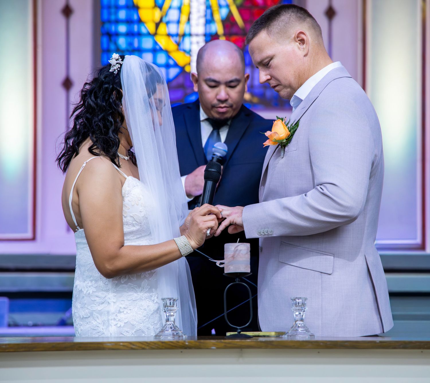 Bride and groom exchanging rings at church altar with officiant and stained glass window backdrop.