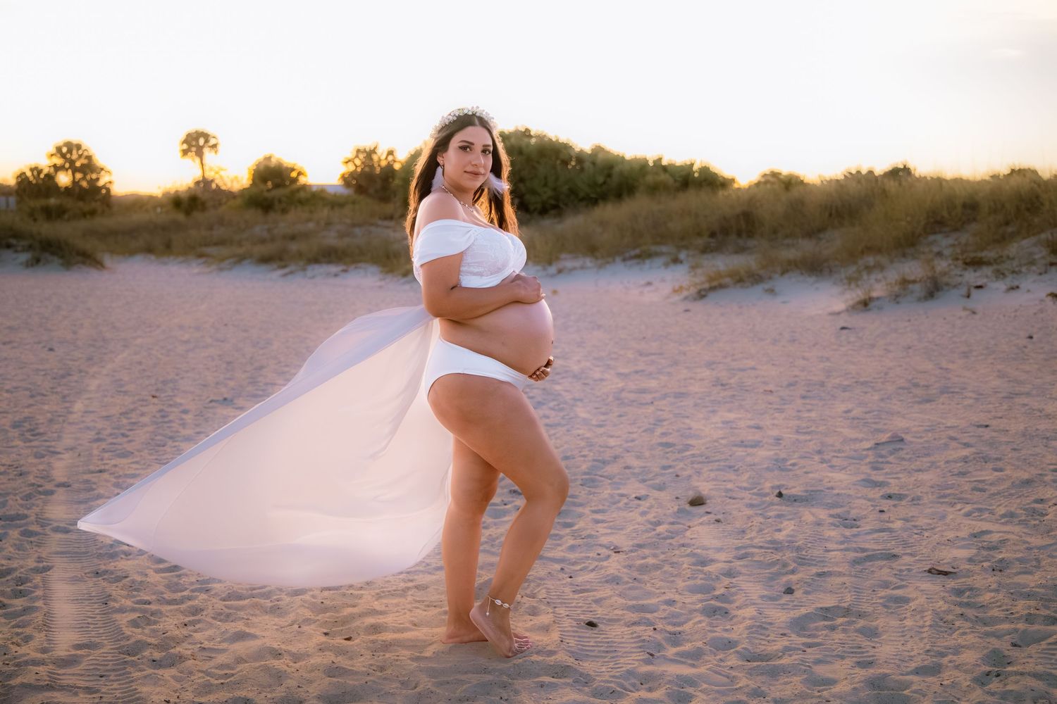Pregnant woman in white bodysuit with flowing skirt poses on sandy beach at golden hour sunset.