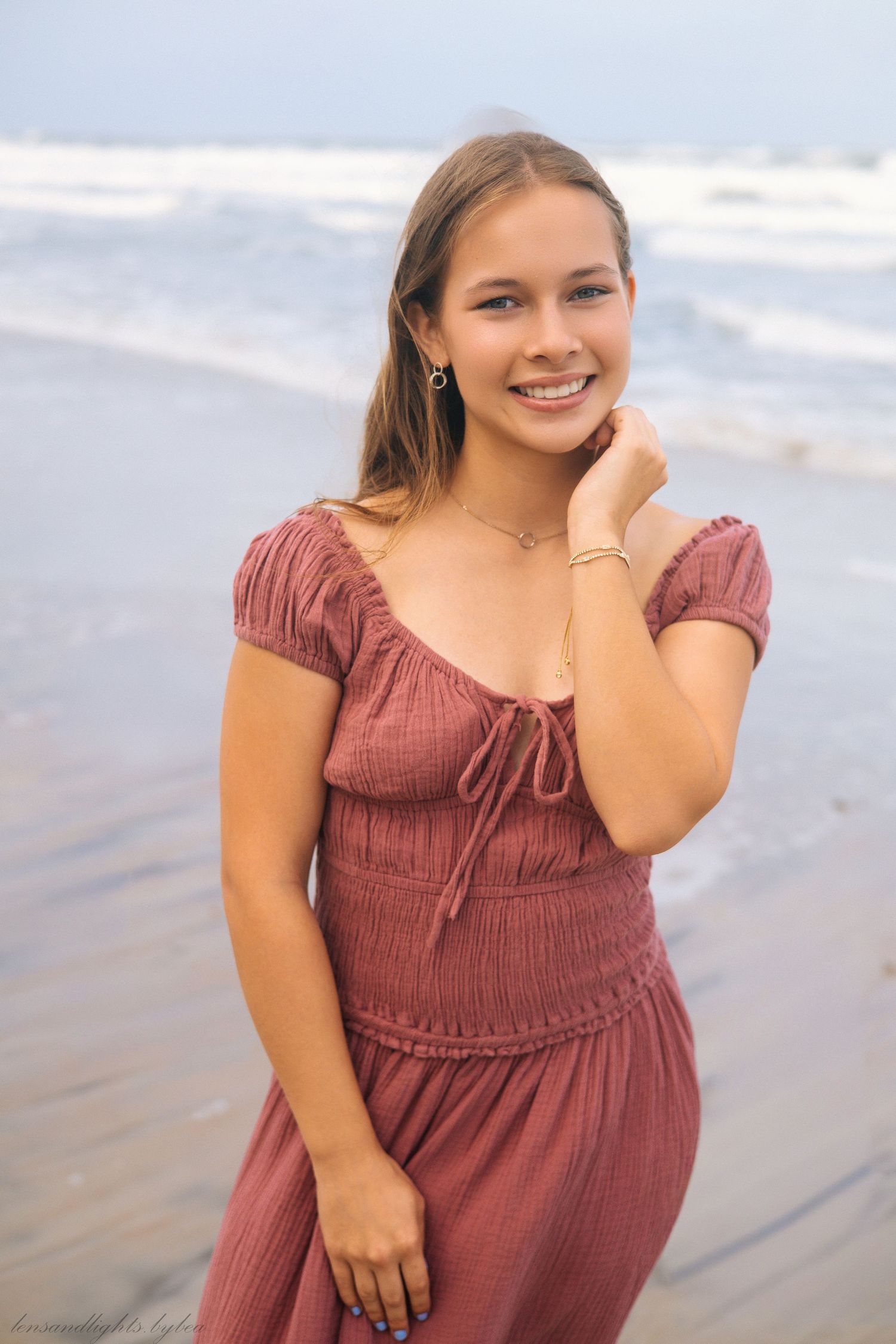 Young woman in a burgundy dress smiles warmly during a beach photoshoot at sunset.