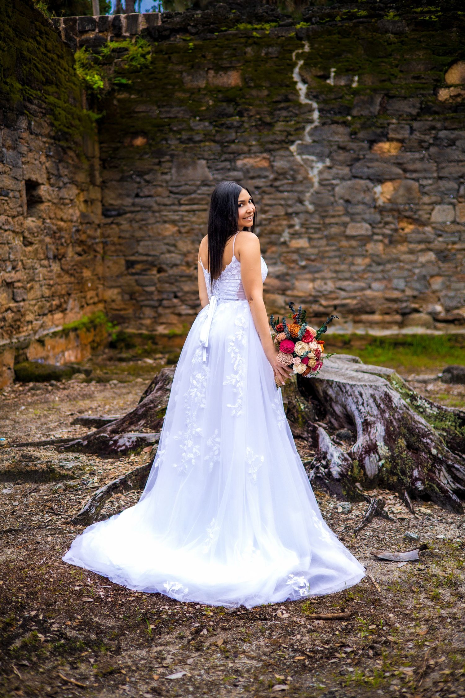 A bride in a flowing white gown sits elegantly on mossy tree roots by a stone wall.