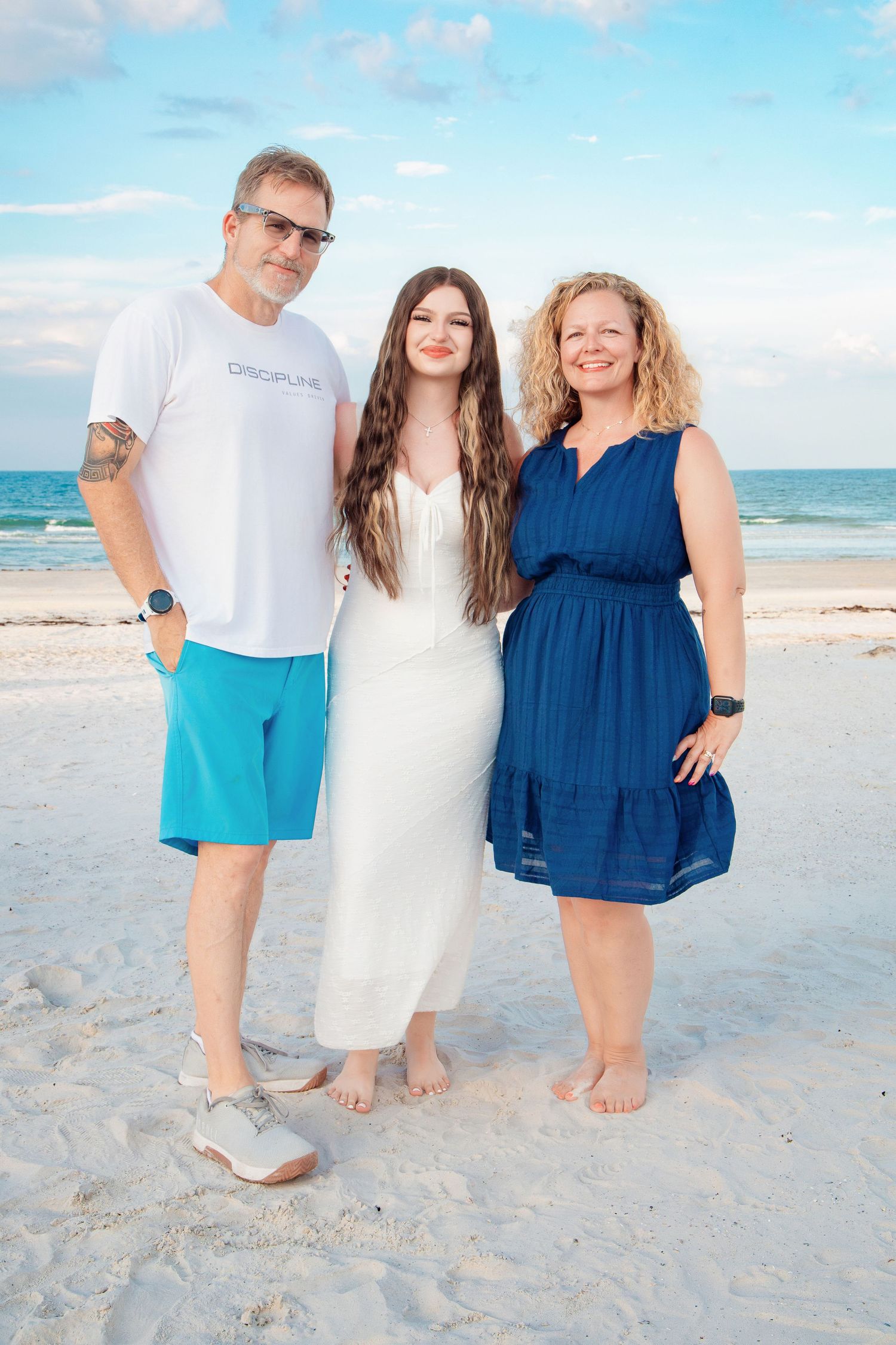 Family of three posing together on a white sand beach, wearing white and blue outfits with ocean waves behind them.