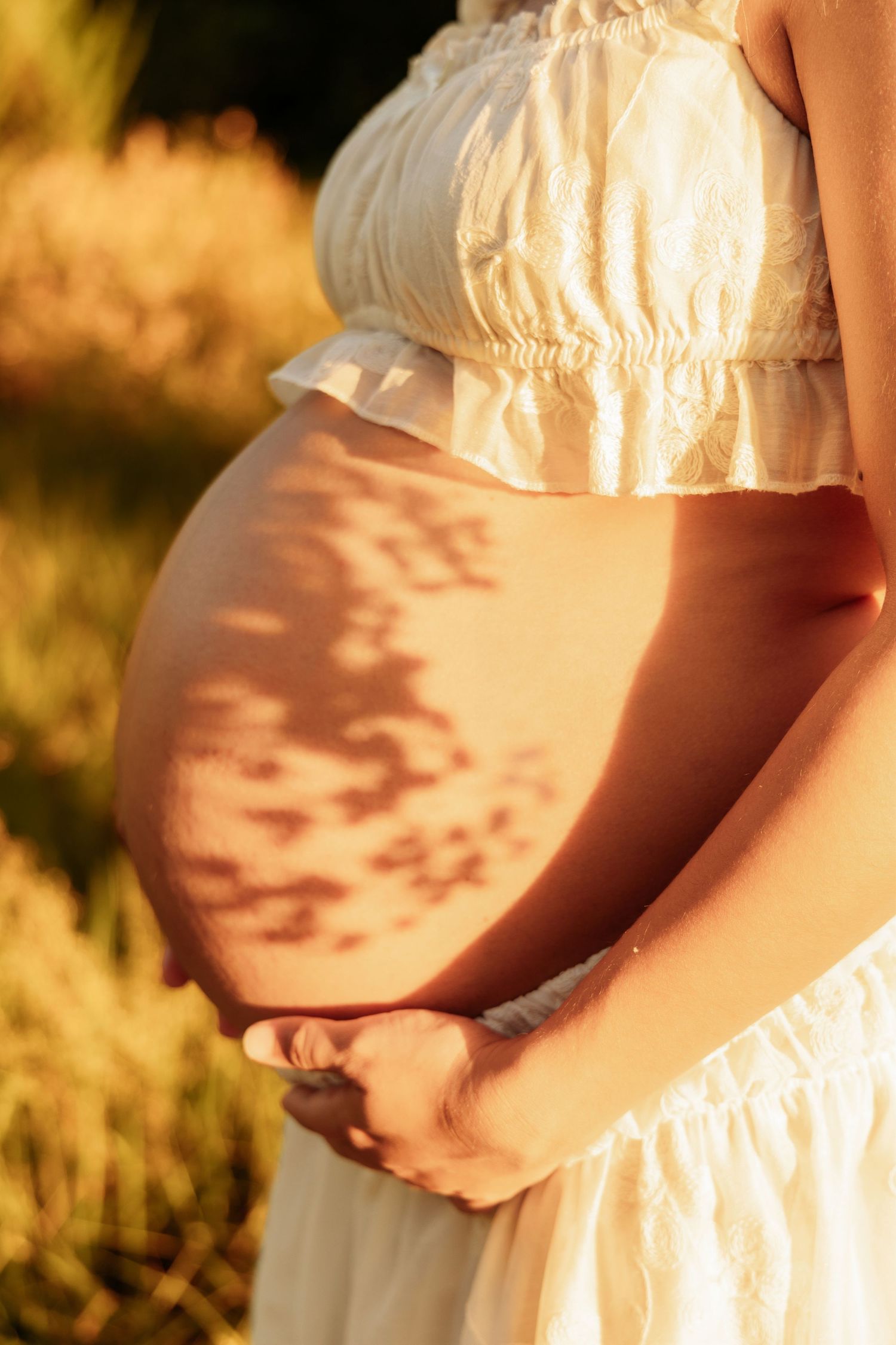 Pregnant woman in white dress cradling bare baby bump outdoors in golden sunlight with dappled shadows.
