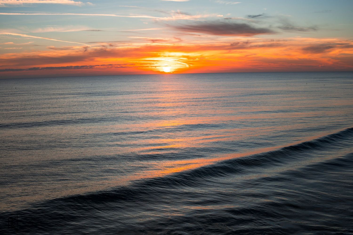 Vibrant orange sunset illuminates the horizon over calm ocean waves with dramatic clouds in the sky.
