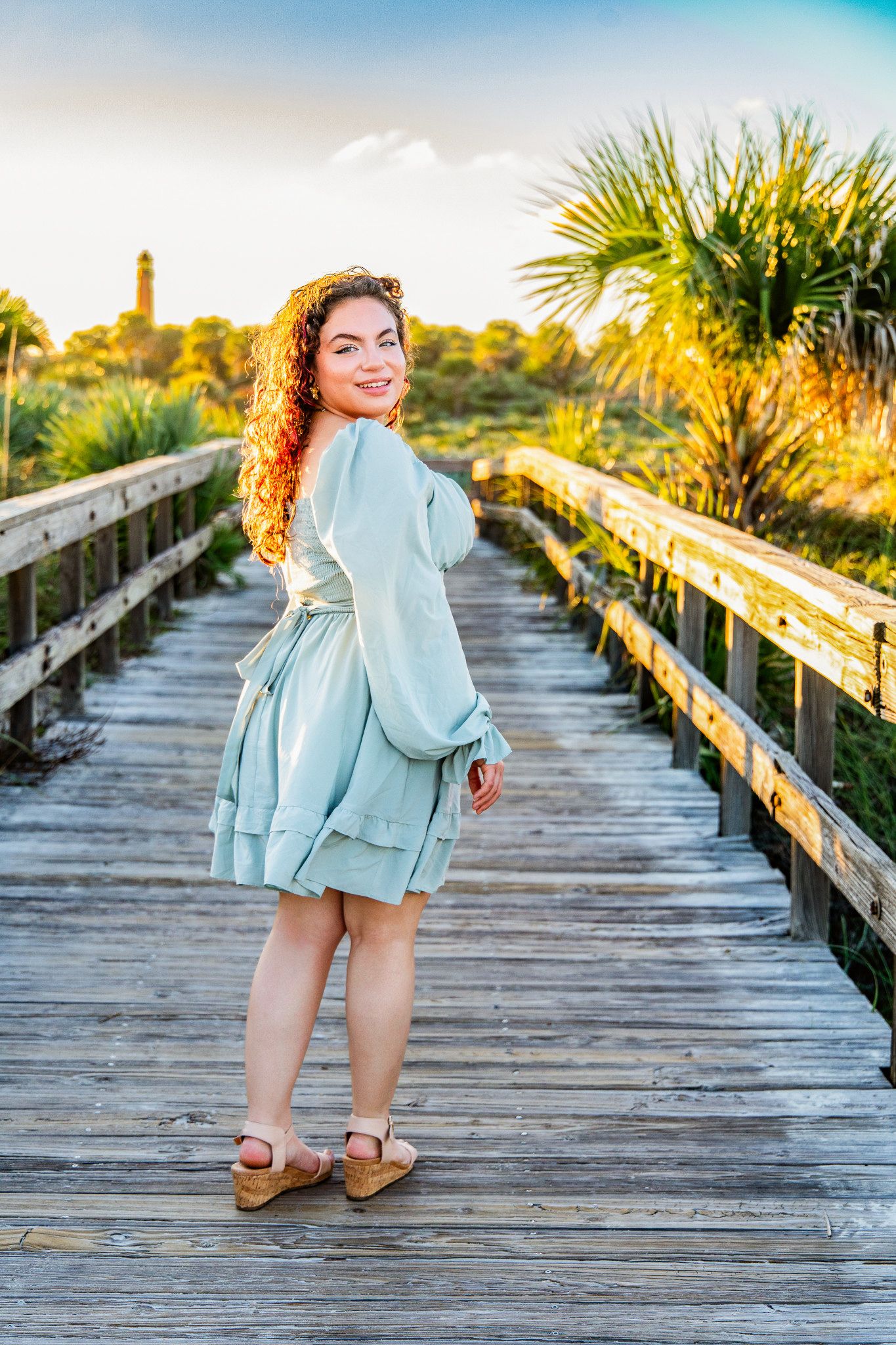 Smiling young woman with curly hair in a mint green dress looks back over her shoulder on a wooden boardwalk at sunset near a lighthouse.