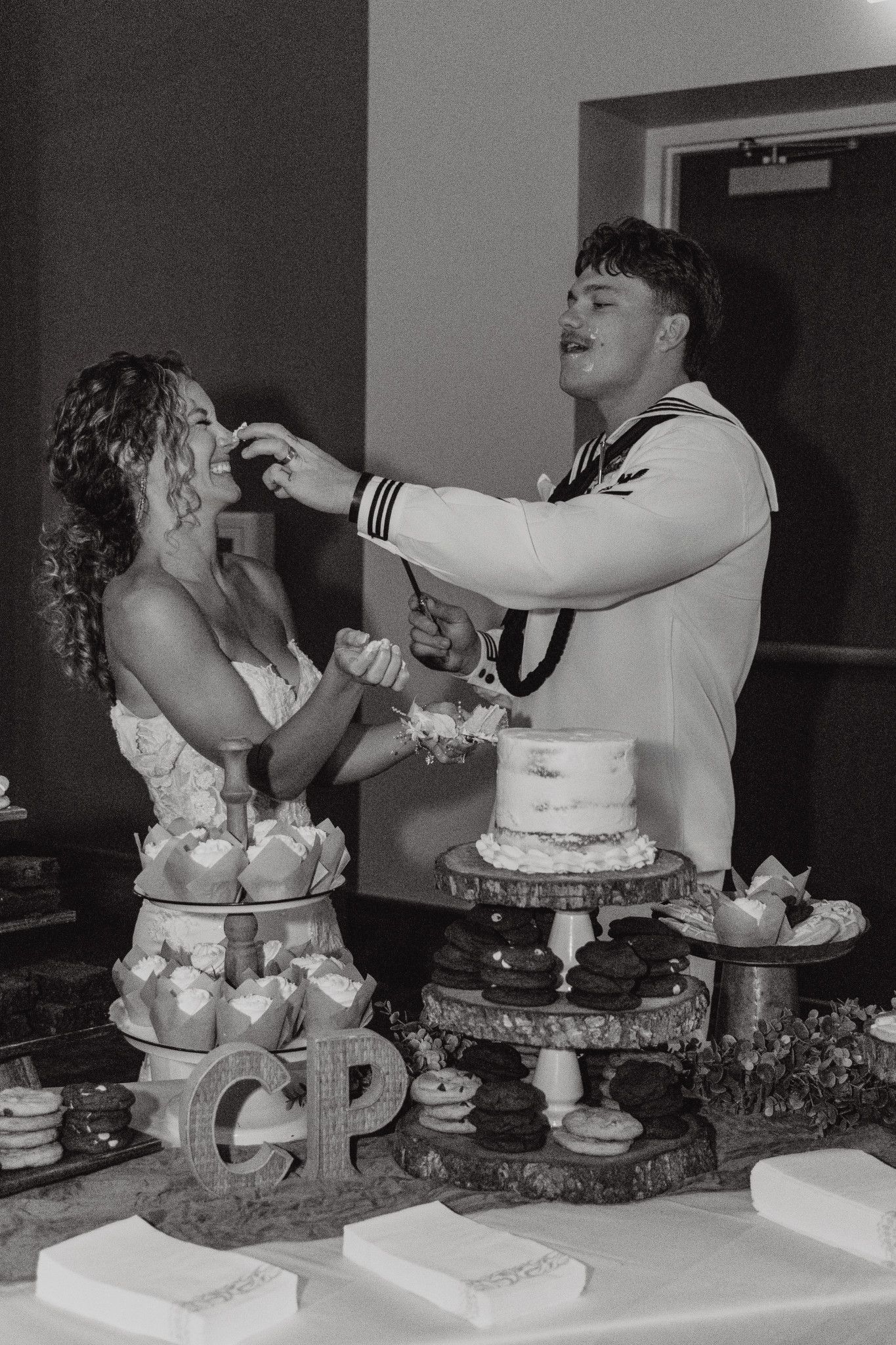 Black and white photo of bride and groom smashing wedding cake, dessert table with monogram CP display.