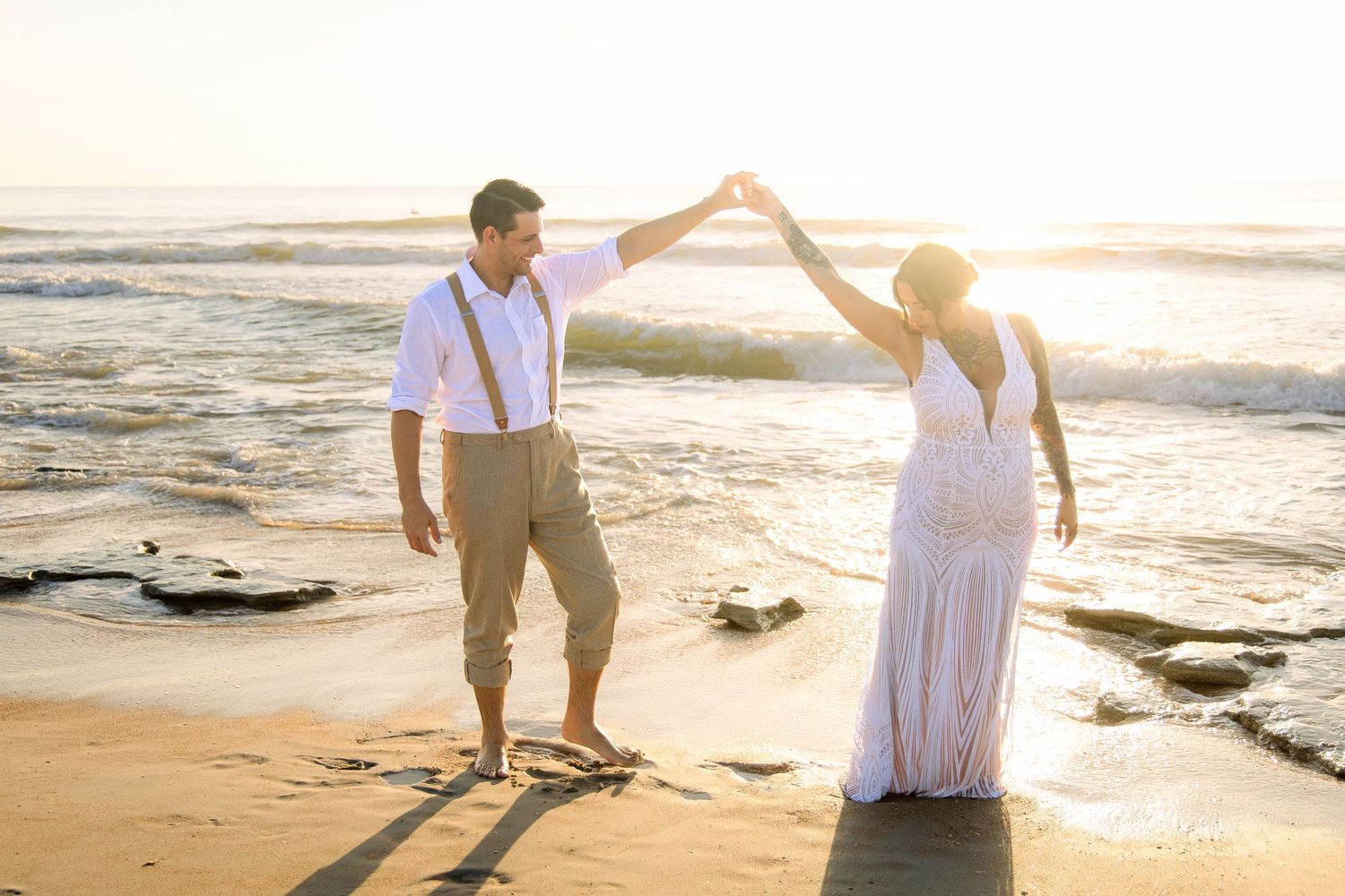 Couple dancing on a sandy beach at golden hour, bride in white dress, groom in suspenders.