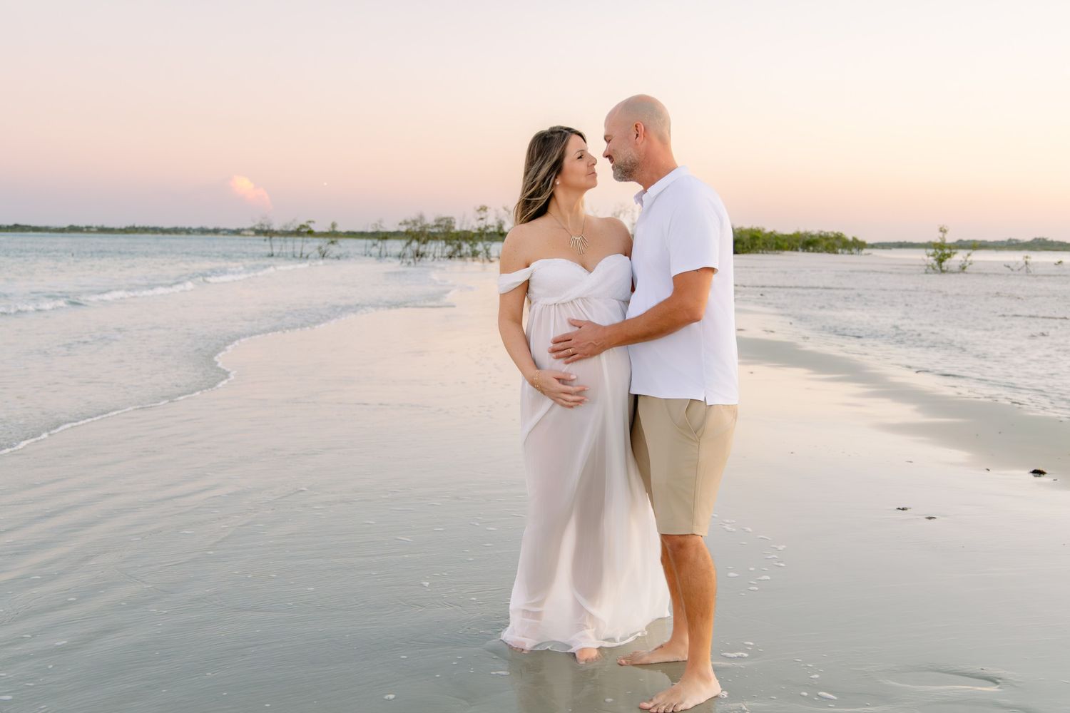 Pregnant couple standing barefoot on a beach at sunset, woman in white dress cradling baby bump.