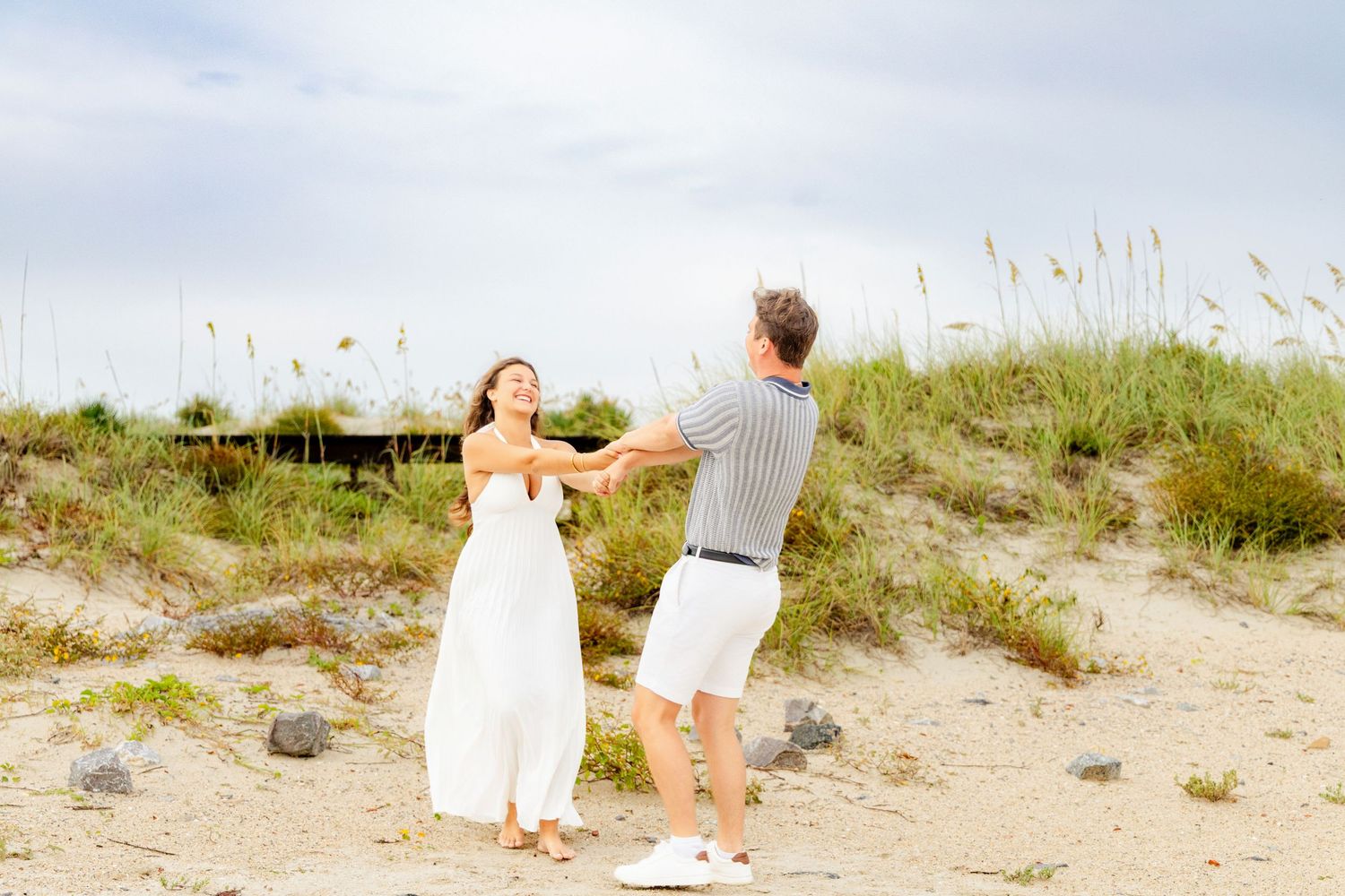 Couple dancing joyfully on a sandy beach near grass dunes on a cloudy day.