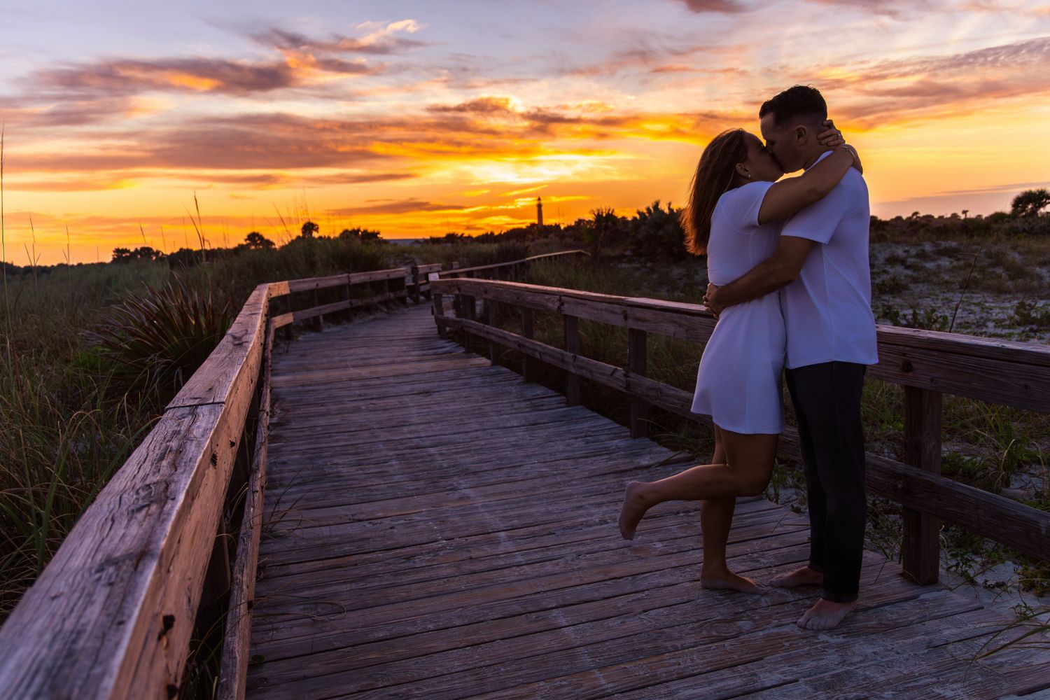 Couple embracing and kissing on a wooden boardwalk at sunset with vibrant orange and purple skies.